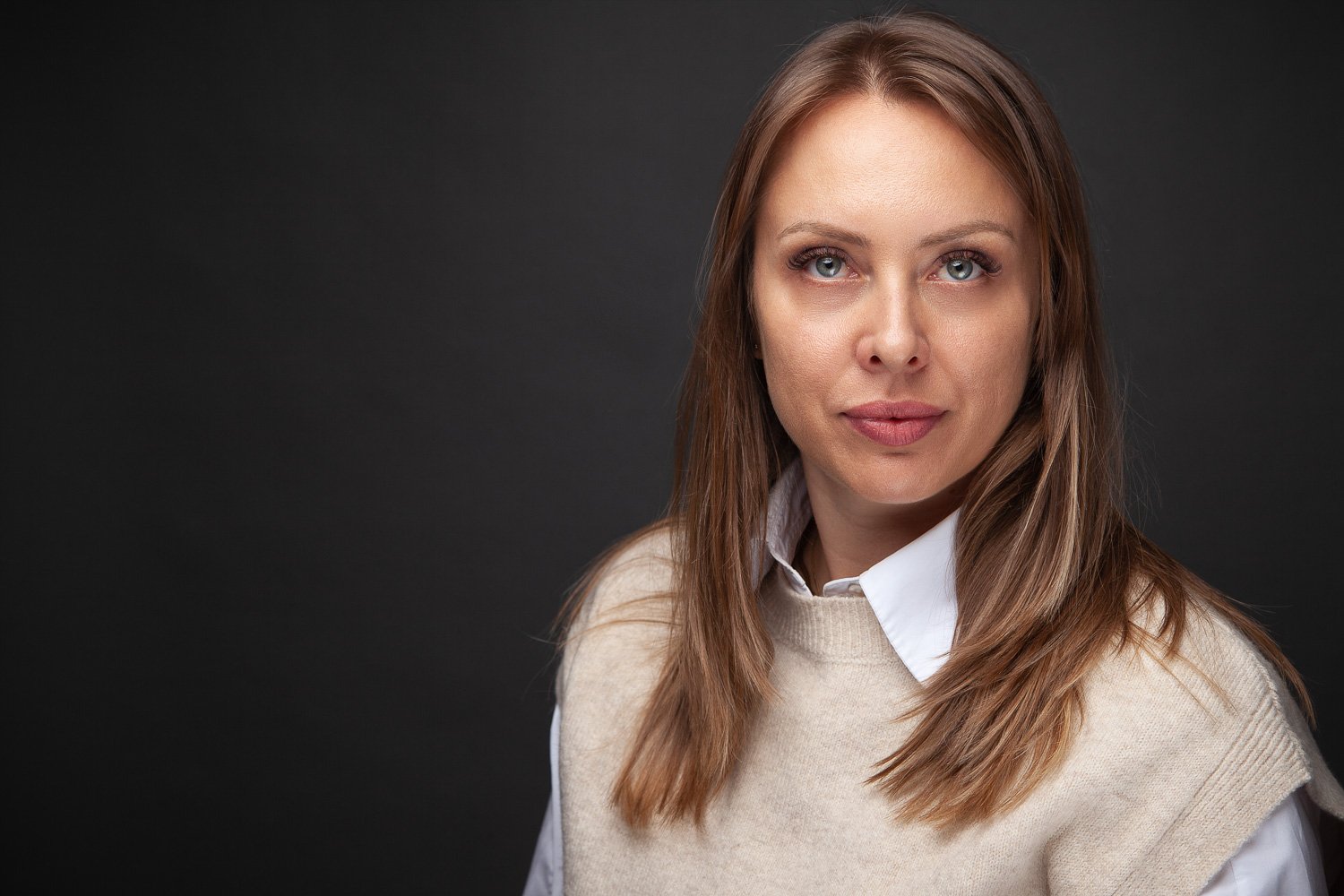 Woman with long wavy brown hair, cream jumper with white collar, professional headshot London against a dark background