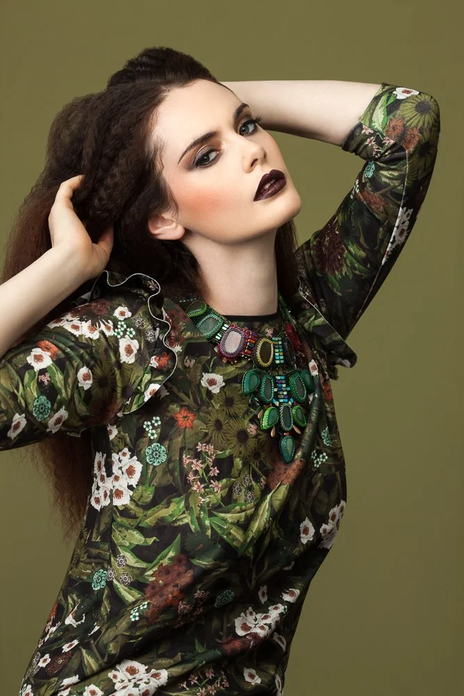 Model with hands in her crimped hair, wearing a floral print top and jewelled necklace, olive backdrop, styled and art directed by Laurence Trolet, branding photography