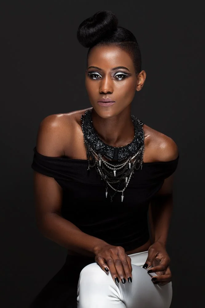 Seated model in an off-shoulder black top and white trousers, wearing a statement bib necklace, styled and art directed by Laurence Trolet, brand photography