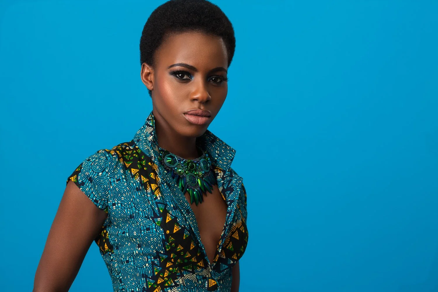Model with short cropped hair in a blue and yellow geometric print top, wearing a statement necklace, blue backdrop, styled and art directed by Laurence Trolet, branding photographer
