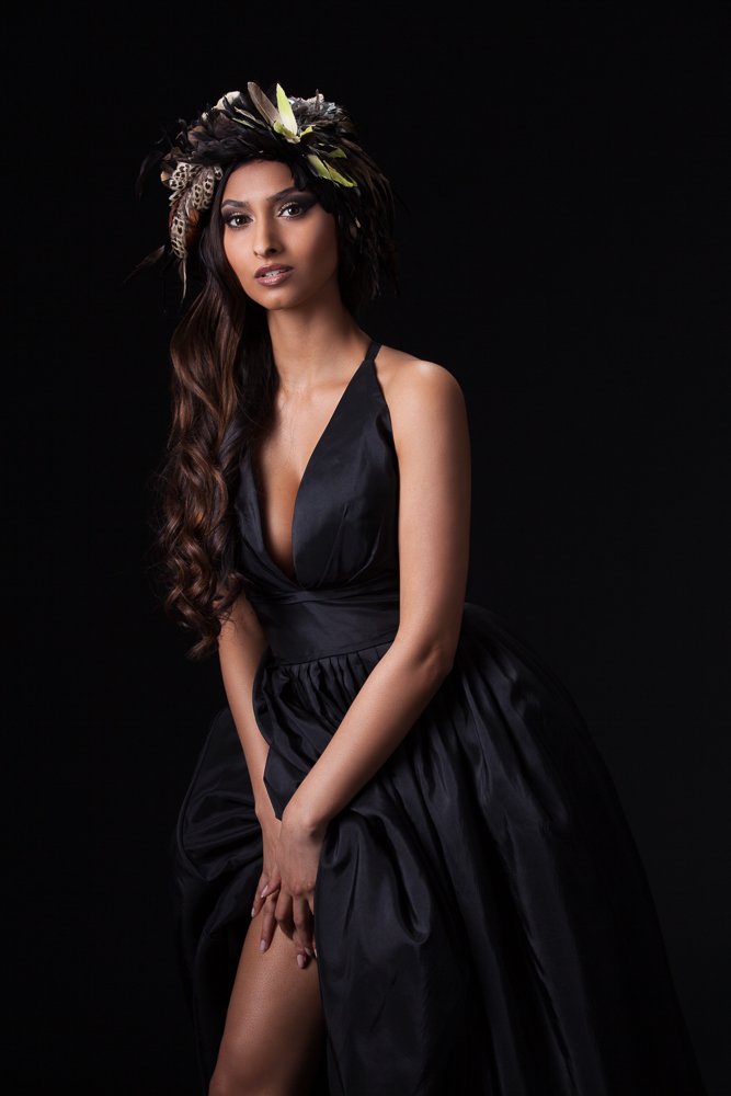 Model in a feathered headpiece and black halter gown with a thigh-high slit, leaning forward against a dark backdrop, styled and art directed by Laurence Trolet, brand photography