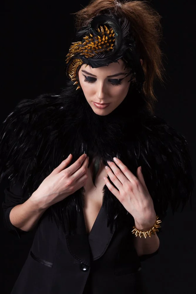 Close-up of a model with lowered eyes in a gold and black feathered headpiece and feather cape, styled and art directed by Laurence Trolet, brand photographer
