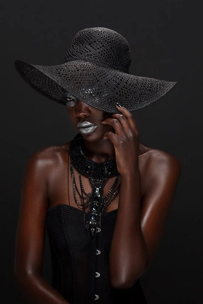 Model in a black wide-brim woven hat and corset, wearing a layered black necklace, styled and art directed by Laurence Trolet, brand photography