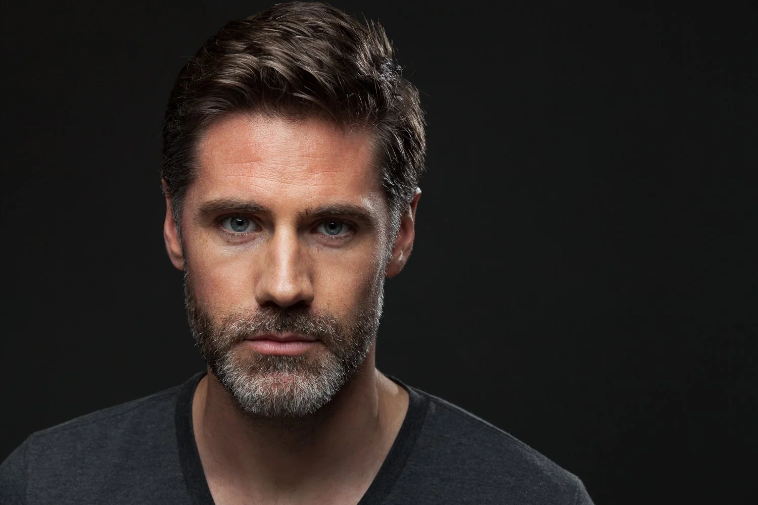 Man with wavy brown hair and a salt and pepper beard, dark t-shirt, moody headshot against a dark background