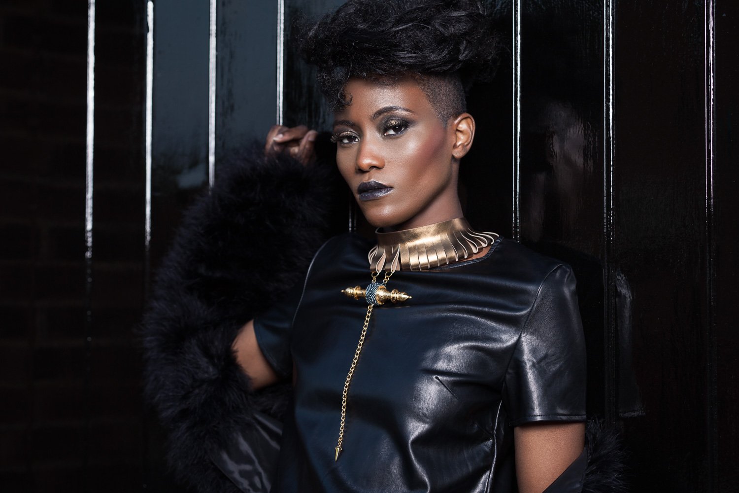 Model in a black leather top with a gold collar necklace and long pendant, posed against dark metal bars, styled and art directed by Laurence Trolet, brand photographer