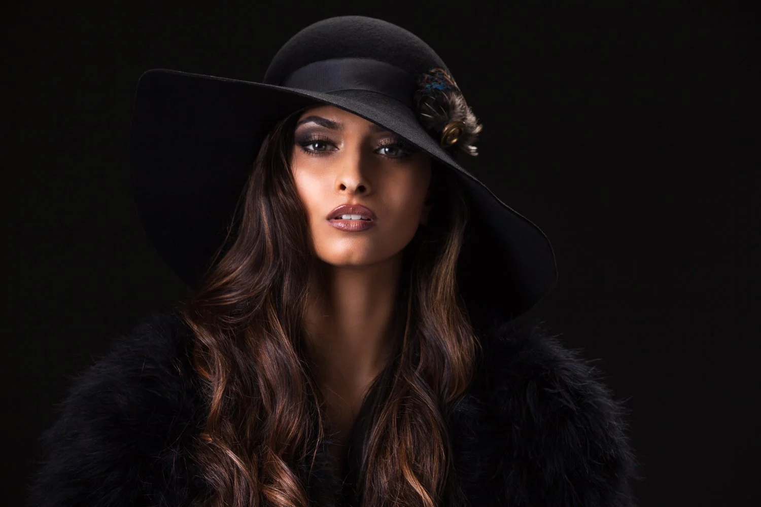Model in a black wide-brim hat with a decorative band detail and a black fluffy jacket, dark backdrop, styled and art directed by Laurence Trolet, brand photography