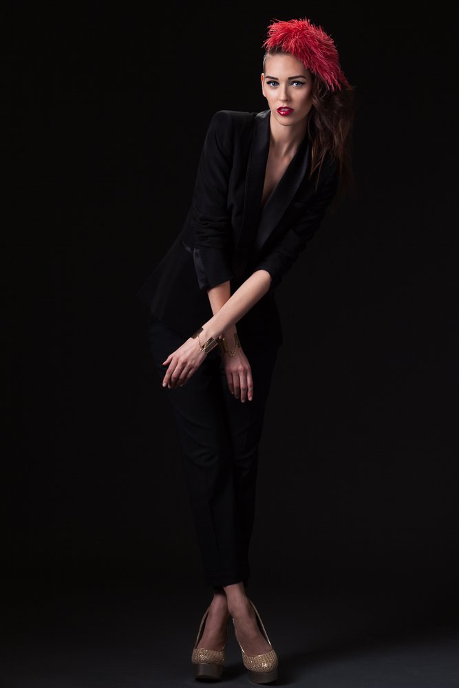 Full-length portrait of a model in a red feathered headpiece and black trouser suit, styled and art directed by Laurence Trolet, branding photographer