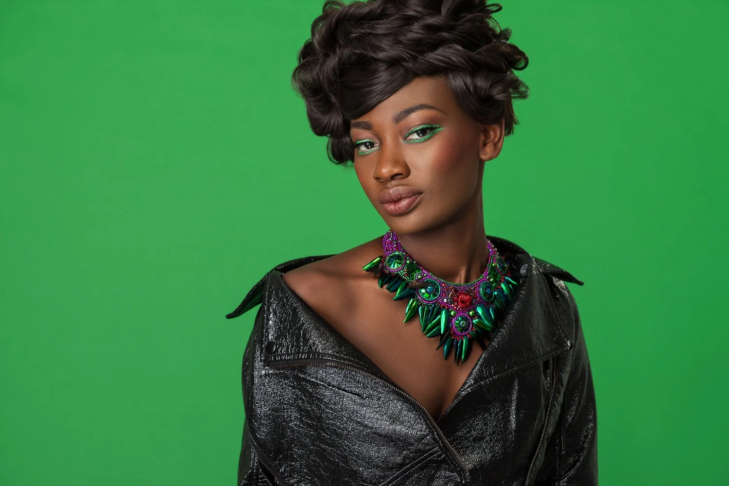 Model with a sculpted curled updo and green eye makeup, black leather jacket and jewelled statement necklace, green backdrop, styled and art directed by Laurence Trolet, brand photographer