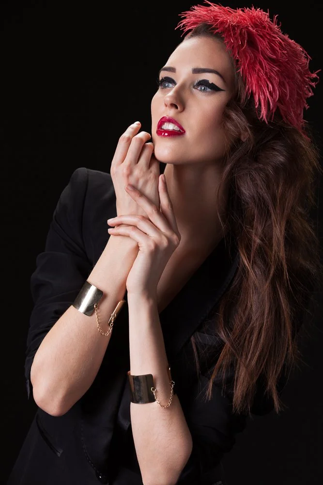 Model in a red feathered headpiece and gold cuff bracelets, hands raised to her face, black jacket, styled and art directed by Laurence Trolet, brand photographer