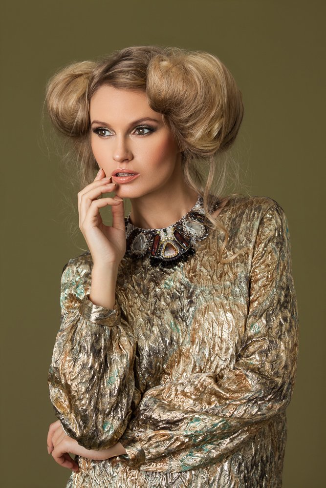 Model with sculpted blonde curls and a hand to her chin, metallic gold top and jewelled necklace, styled and art directed by Laurence Trolet, brand photographer