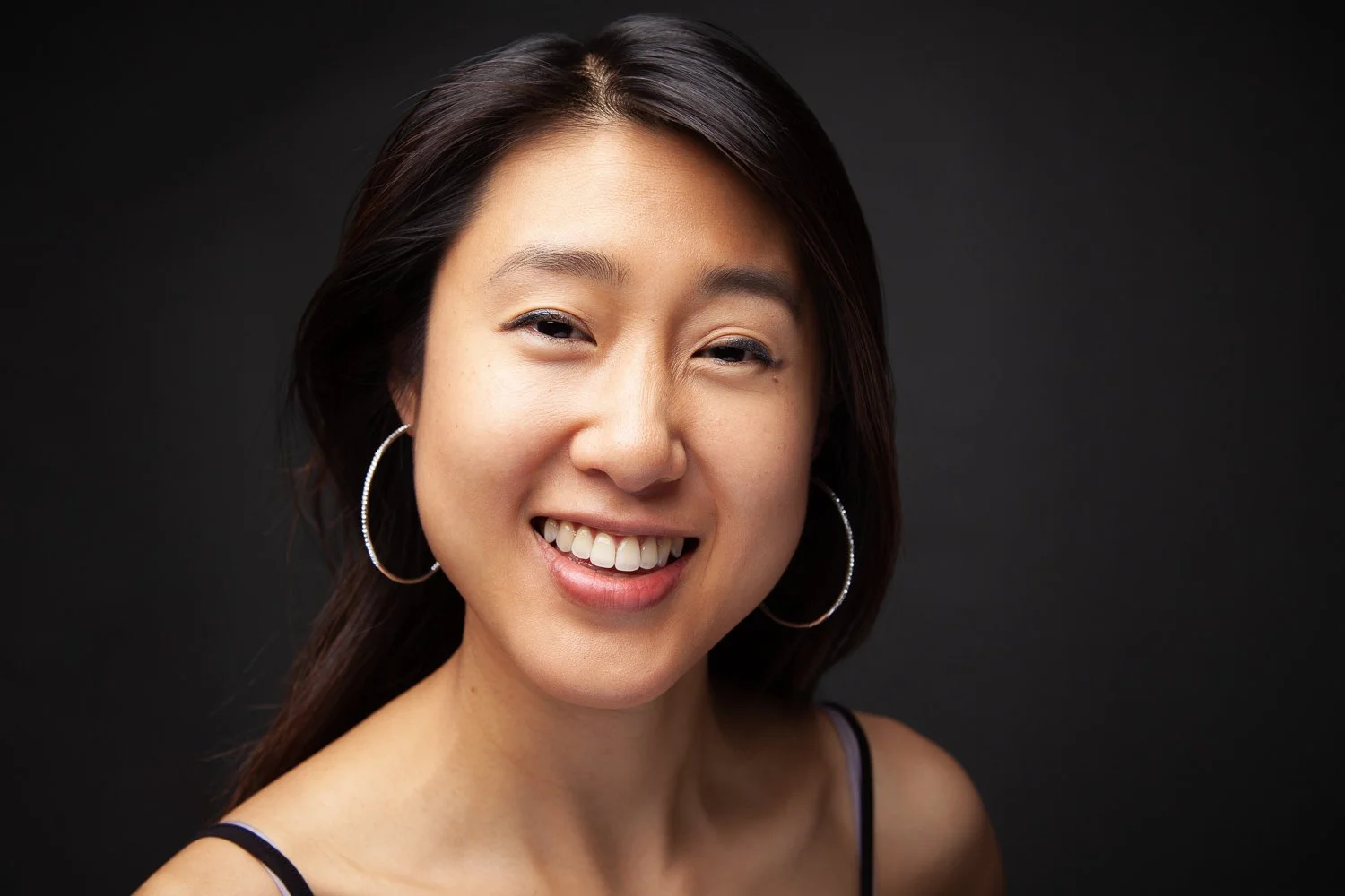 Woman with sleek dark hair, hoop earrings, smiling headshot against a grey background