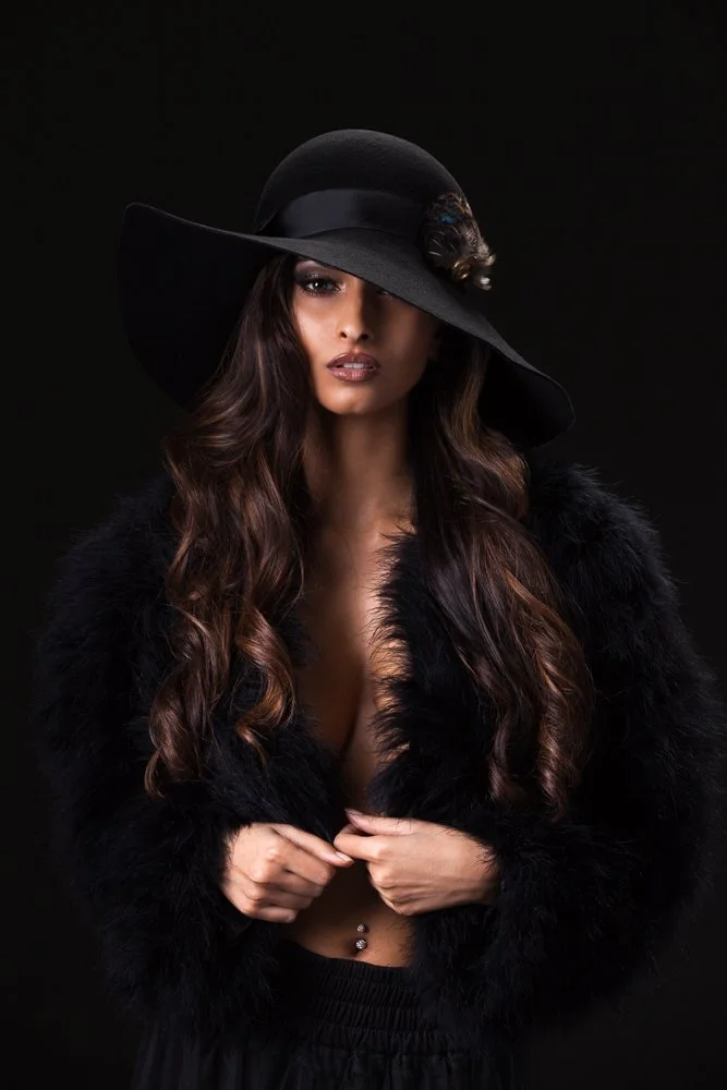 Model in a black wide-brim hat with long wavy hair, holding a black fluffy jacket closed, styled and art directed by Laurence Trolet, branding photographer