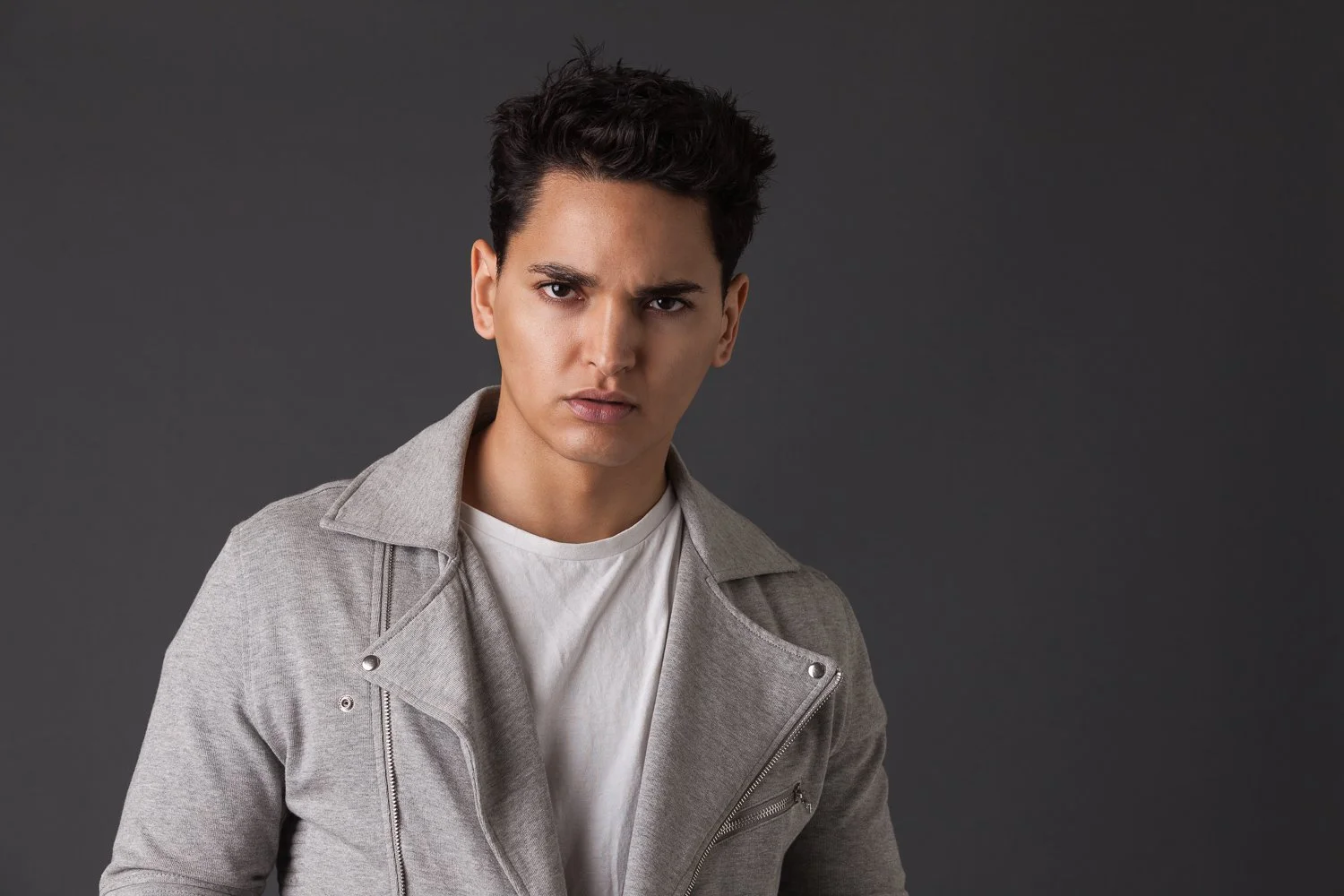 Young man with short hair, grey jacket, intense expression against a grey background