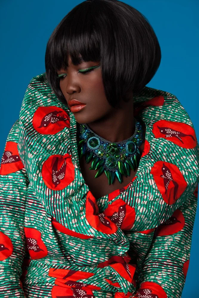 Model with a sleek black bob in a red and green print top, wearing a green statement necklace, blue backdrop, styled and art directed by Laurence Trolet, brand photographer