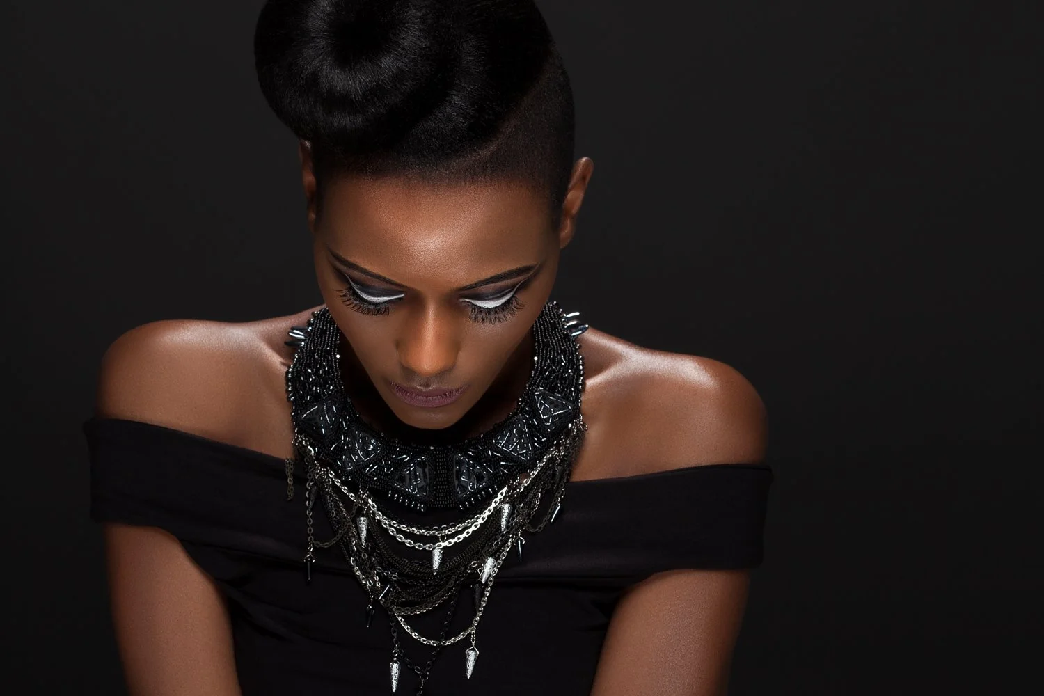 Model with lowered gaze in an off-shoulder black top, wearing a beaded bib necklace with silver chains, styled and art directed by Laurence Trolet, branding photographer