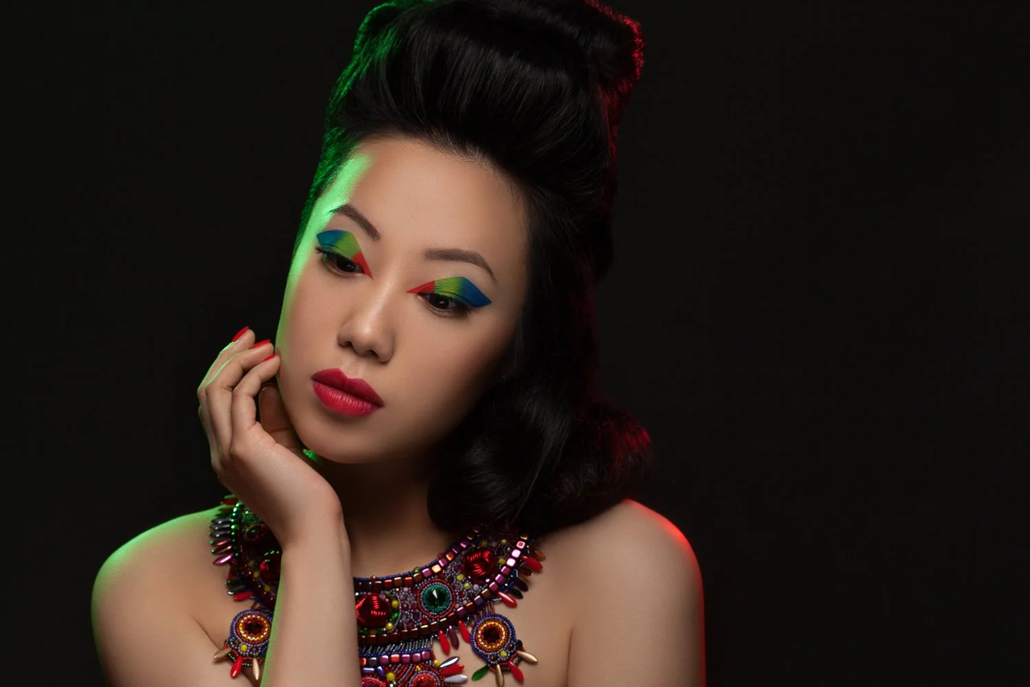 Model touching her face with lowered eyes under coloured rim light, wearing an ornate beaded necklace, styled and art directed by Laurence Trolet, brand photographer