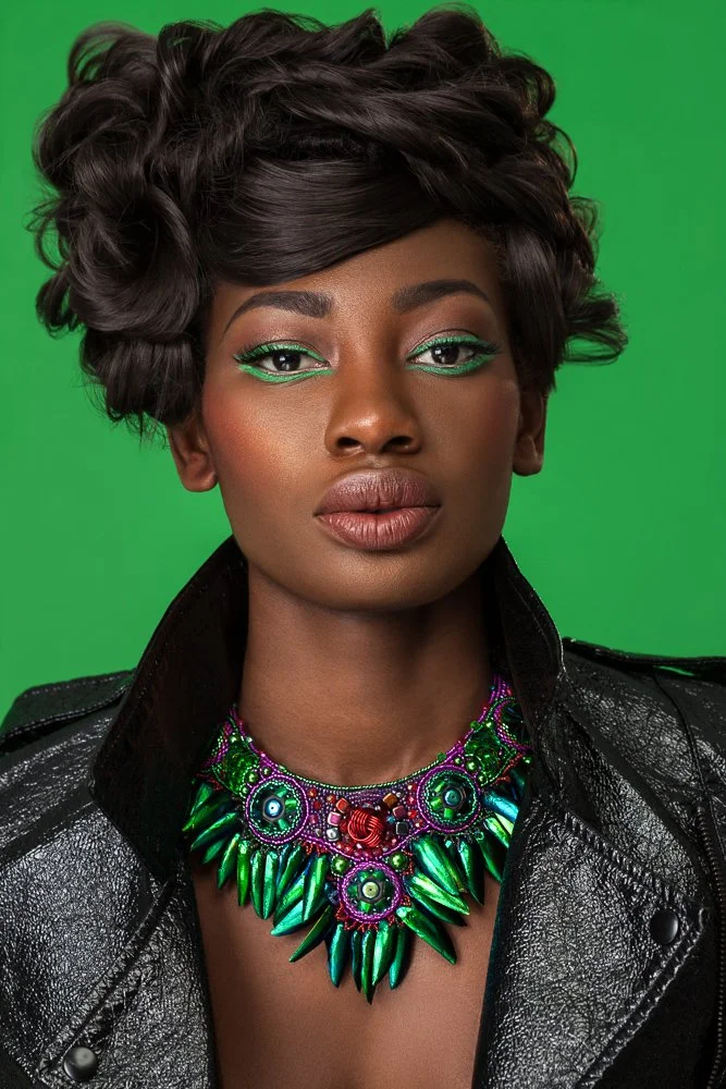 Close-up of a model in a black leather jacket wearing a green and purple jewelled statement necklace, green backdrop, styled and art directed by Laurence Trolet, brand photography