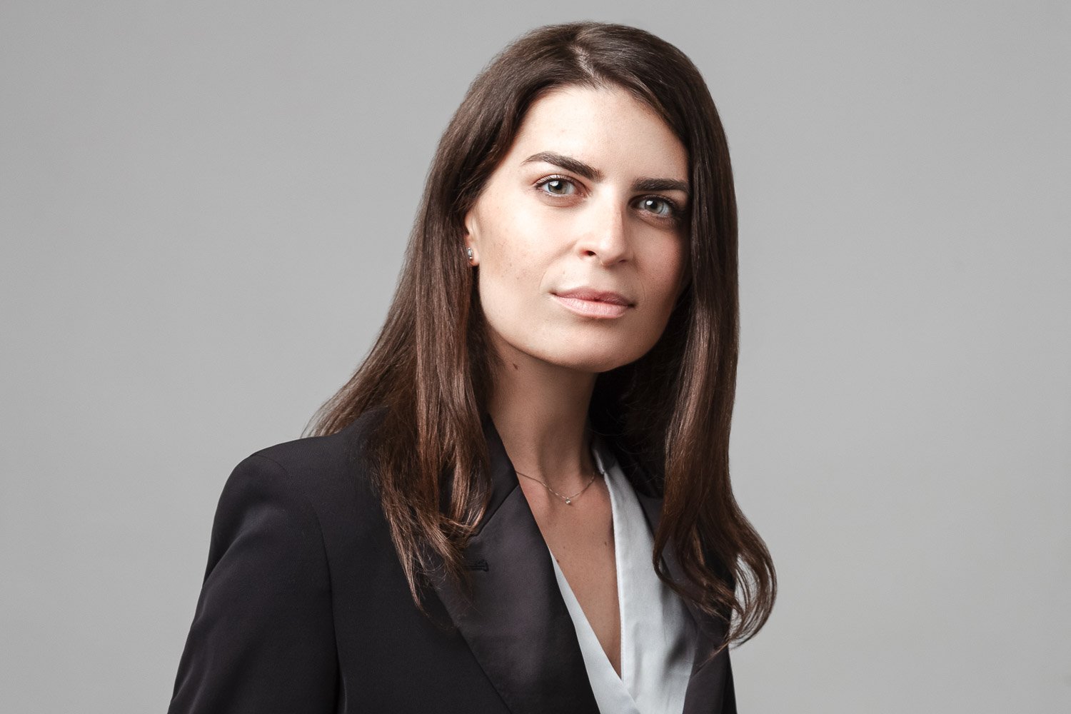 Woman with long dark hair, black blazer, corporate headshot London against a grey background