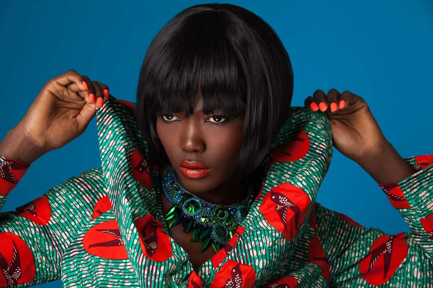 Model with a sleek black bob raising the collar of a red and green print top, green statement necklace, styled and art directed by Laurence Trolet, brand photography