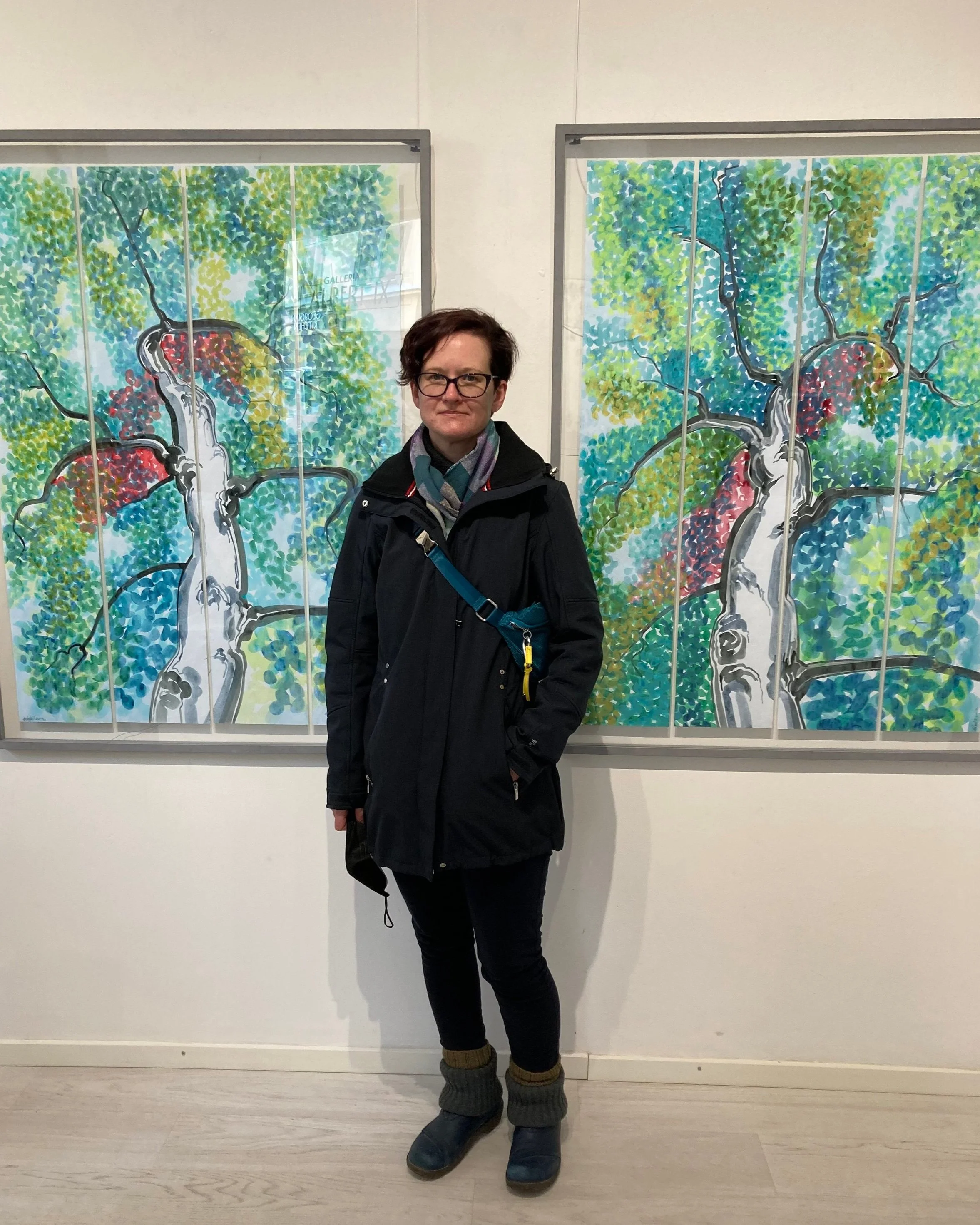 The artist, a middle-aged woman with short brown hair and dressed in very dark blue (coat and jeans, glasses), standing in front of and between the two acrylic paintings hanging on the white gallery wall. She looks bit tired and stony-faced.