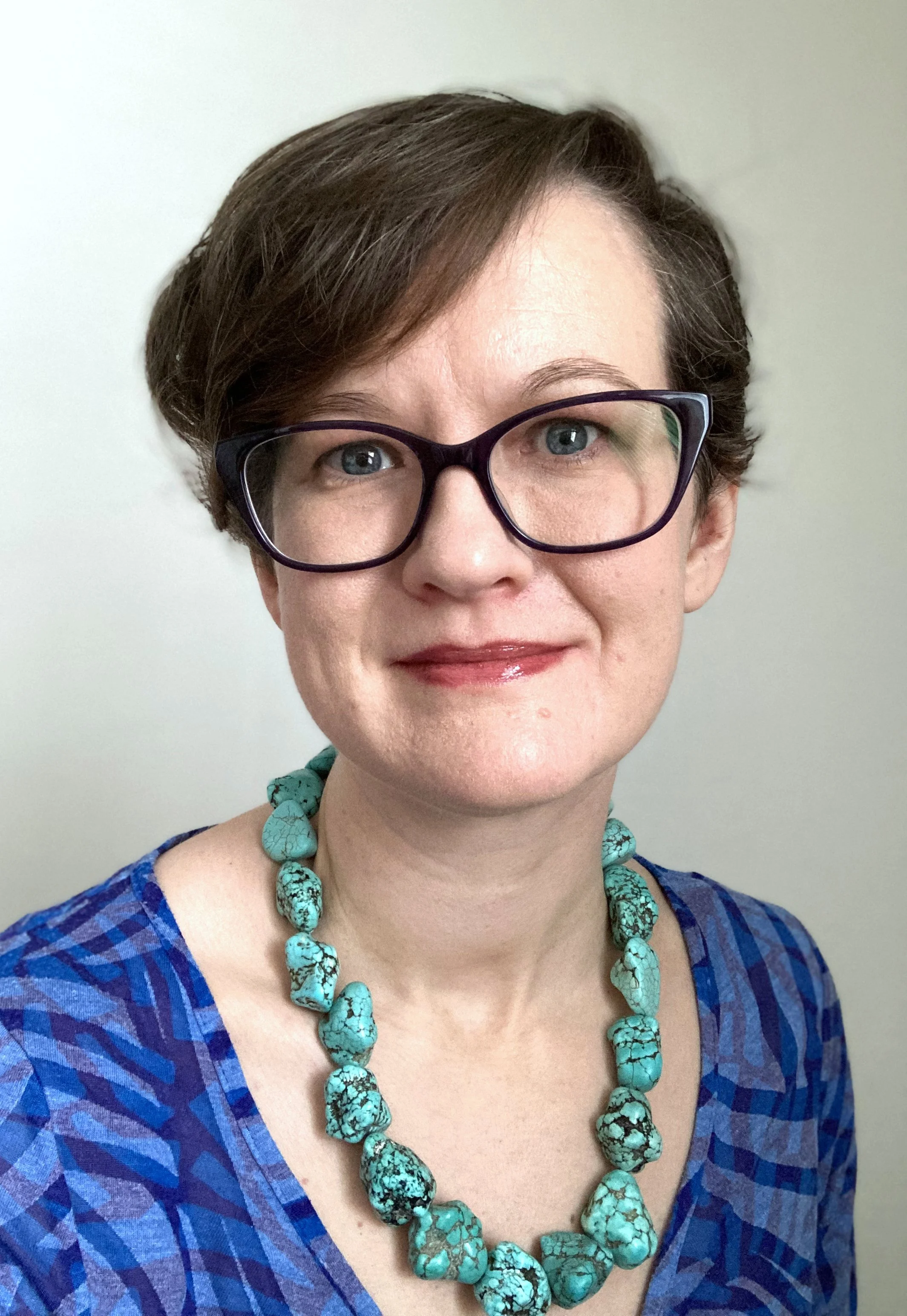 A woman with short brown hair, glasses, and turquoise turquoise beaded necklace wearing a blue patterned top, smiling against a plain background.