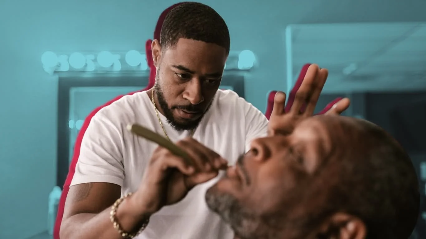 A man with a serious expression performs tattooing on another man's face in a tattoo studio.