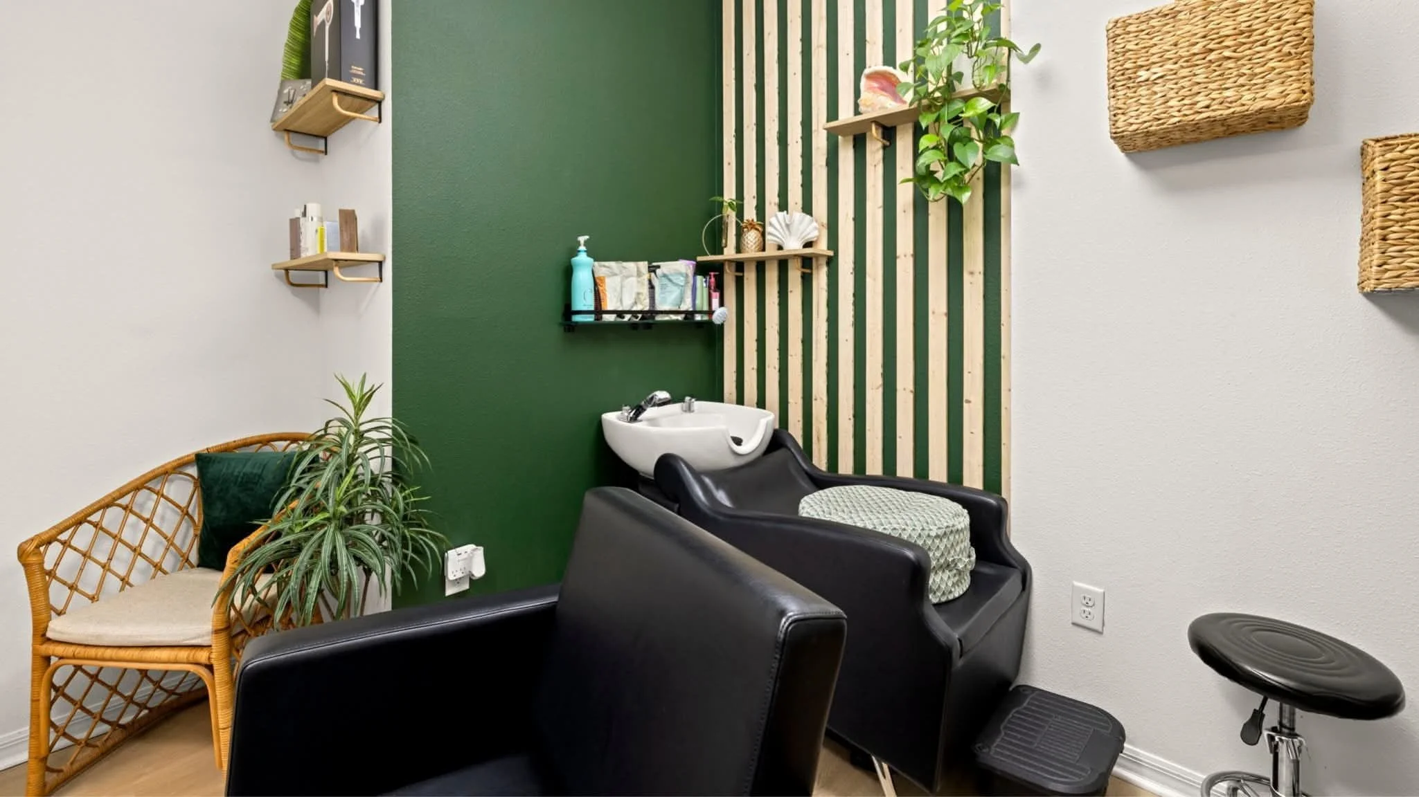 Interior of a hair salon or spa with a black shampoo chair and sink, a woven wicker bench with a green pillow, various wall shelves with decor and products, potted plants, and woven storage baskets on the wall.