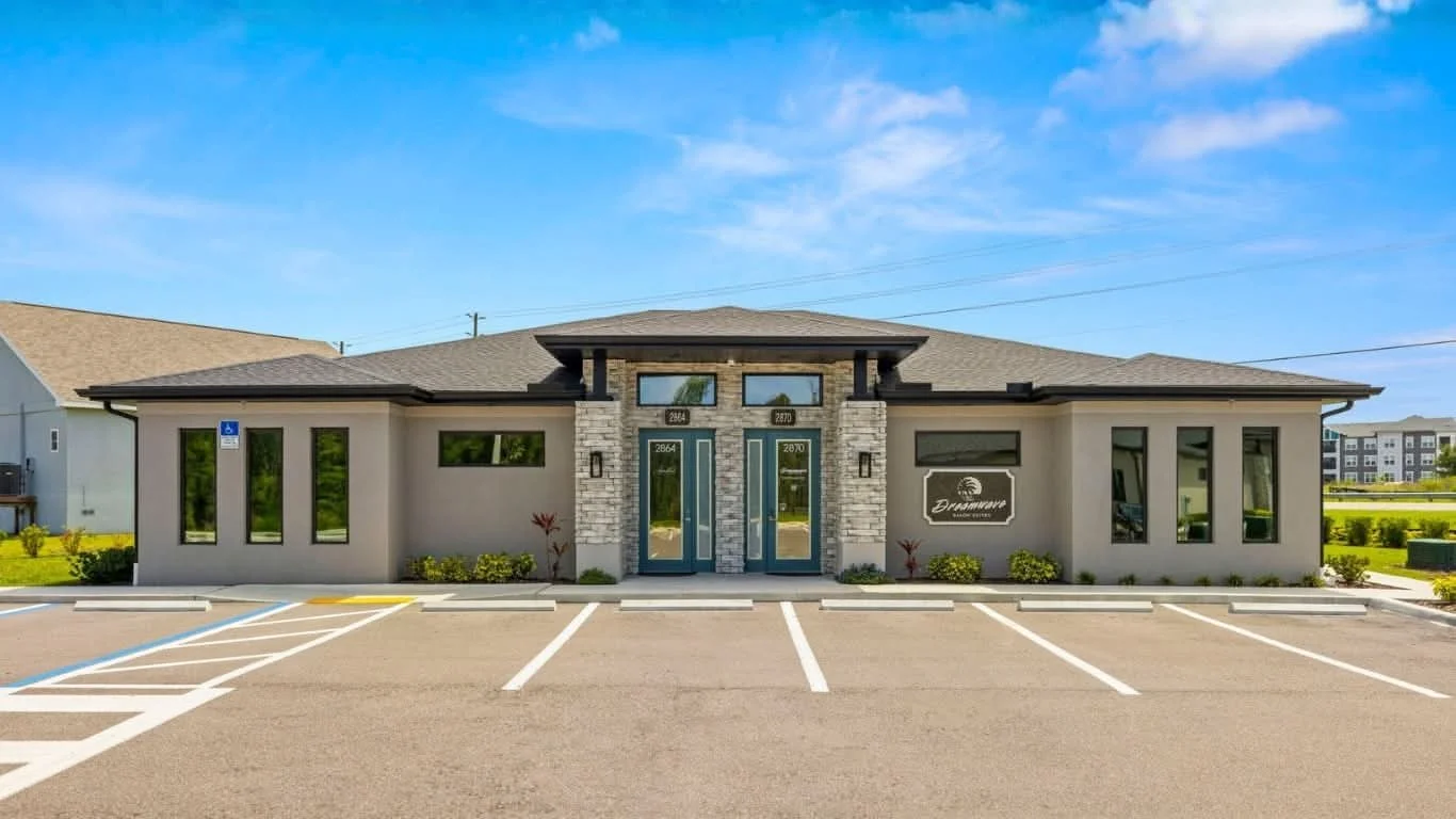 A commercial building with a modern design, featuring a gray brick entrance, multiple windows, and a sign that reads "Dreamscape" in a parking lot with marked spaces, surrounded by green grass and blue skies.