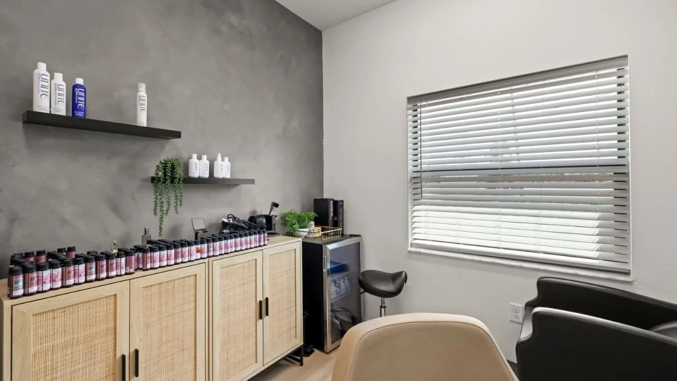 A room with a gray textured wall, a window with horizontal blinds, a black couch, a beige chair, a cabinet with shelves and bottles, and a small refrigerator with plants on top.