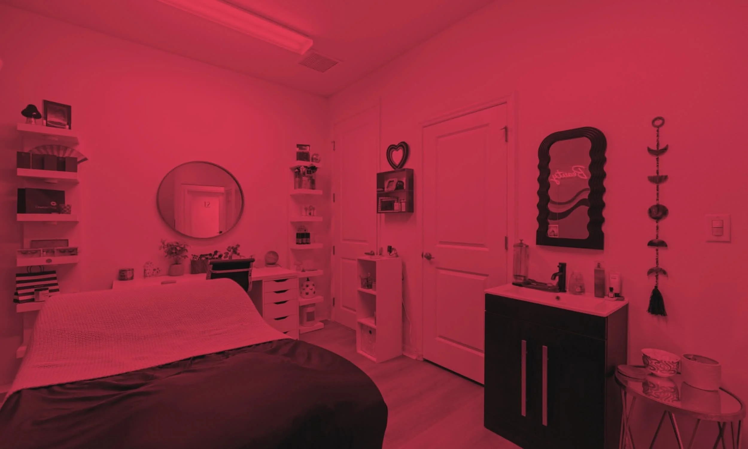 A cozy bedroom with pink lighting. A bed with a blanket is in the foreground, right of the center. Shelves and a round mirror are on the wall behind the bed. There are two white doors, one with the number 12, in the background. A small black cabinet with bottles and a soap dispenser is on the right, next to a small table with toiletries.