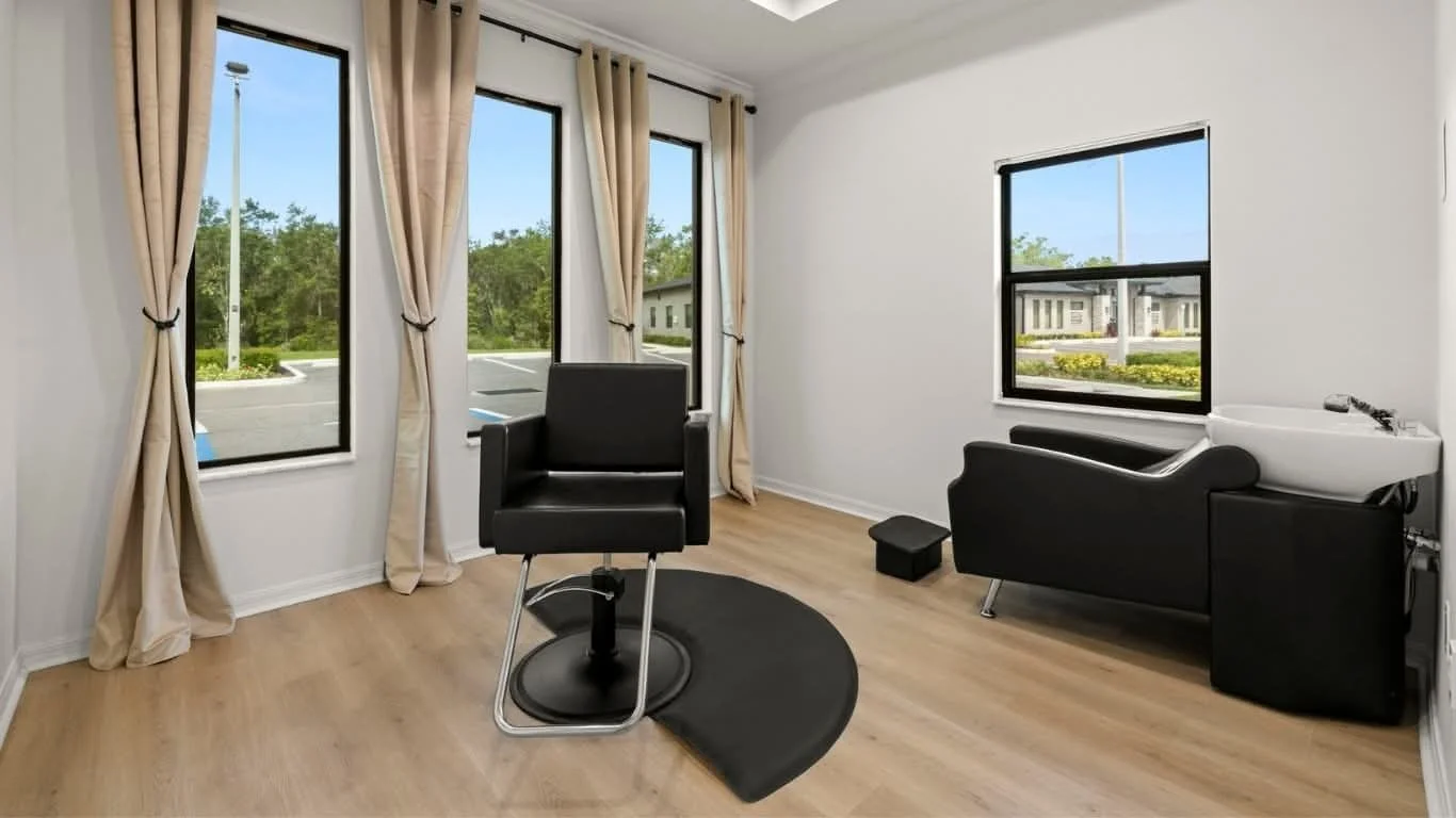 Empty beauty salon room with salon chair, washbasin, and large windows with curtains.