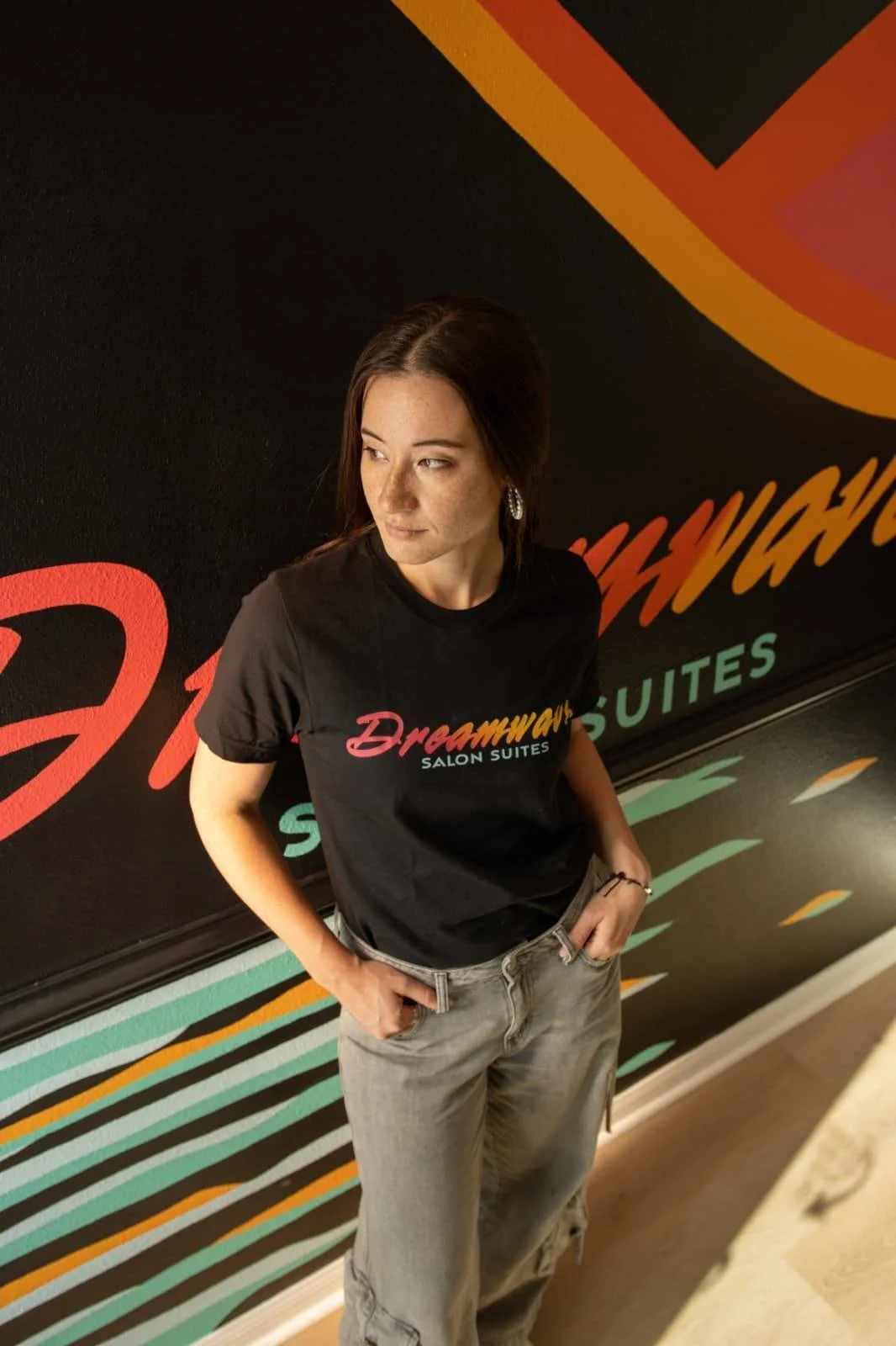 A woman with dark hair, wearing a black t-shirt with the logo 'Dreamway SALON SUITES' and light-colored jeans, standing in front of a colorful wall with the same logo.