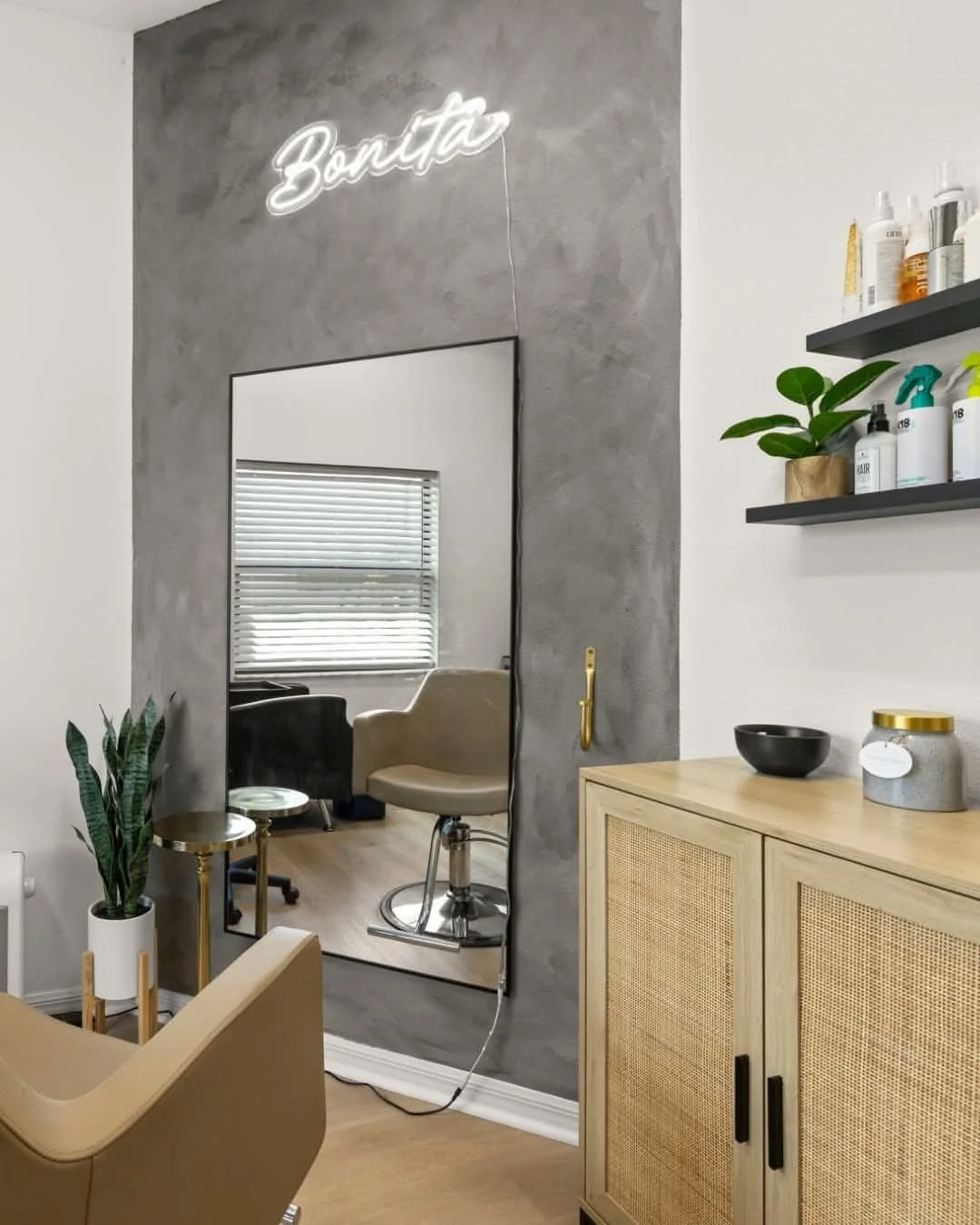 A room with a large mirror on a textured gray wall, reflecting a window with blinds and a beige salon chair. The room features a wooden cabinet, a shelf with hair products, a plant, and a neon sign that says 'Beauty' in cursive.