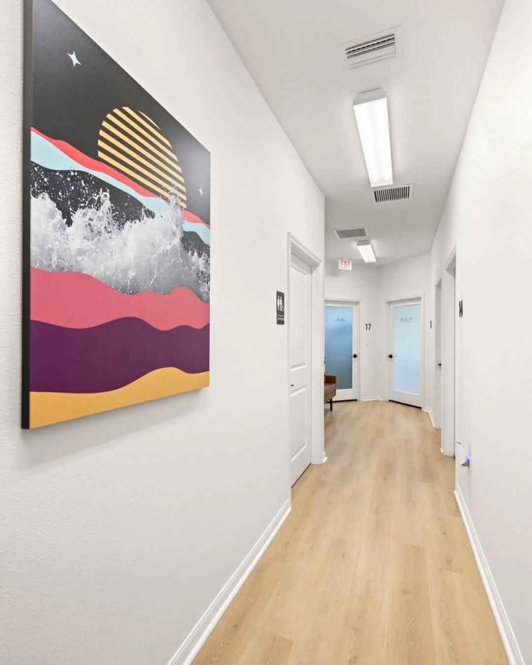 Empty hallway with white walls, light wood flooring, fluorescent lighting, and three doors with frosted glass panels, one with a visible room number 17, and a colorful abstract painting on the left wall.