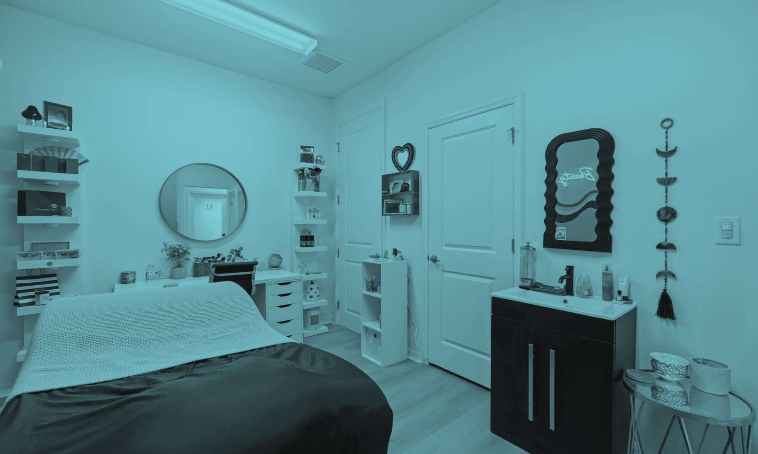 A bedroom with white walls, a bed with a black comforter and white blanket, small bookshelves with decor and framed photos, a circular mirror on the wall, a black dresser with a mirror, and decorative wall hanging.