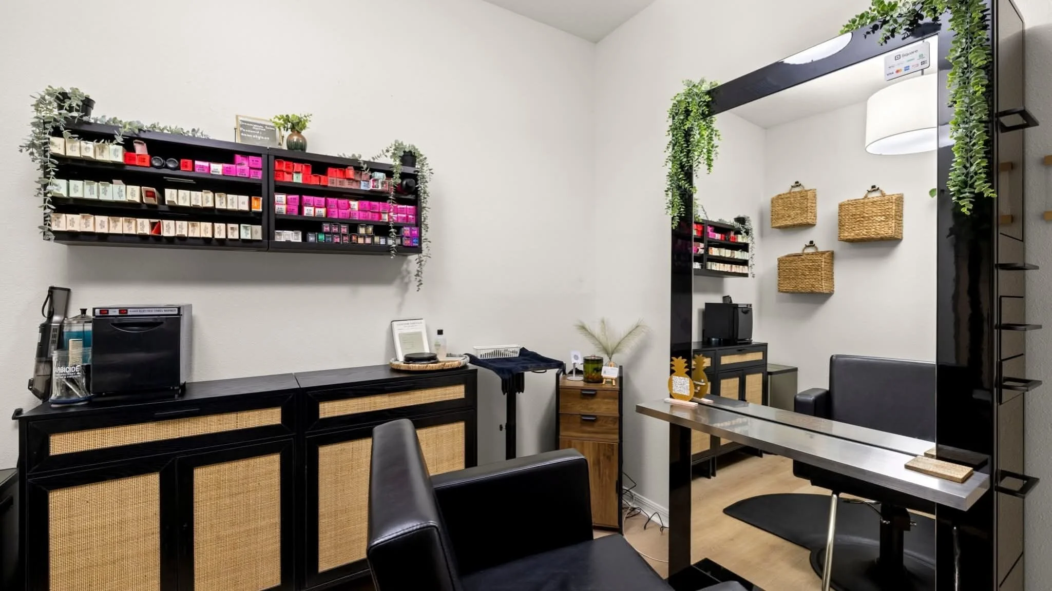 Interior of a modern salon or spa with black and rattan furniture, wall-mounted shelves with colorful product boxes, a large mirror, and decorative greenery.