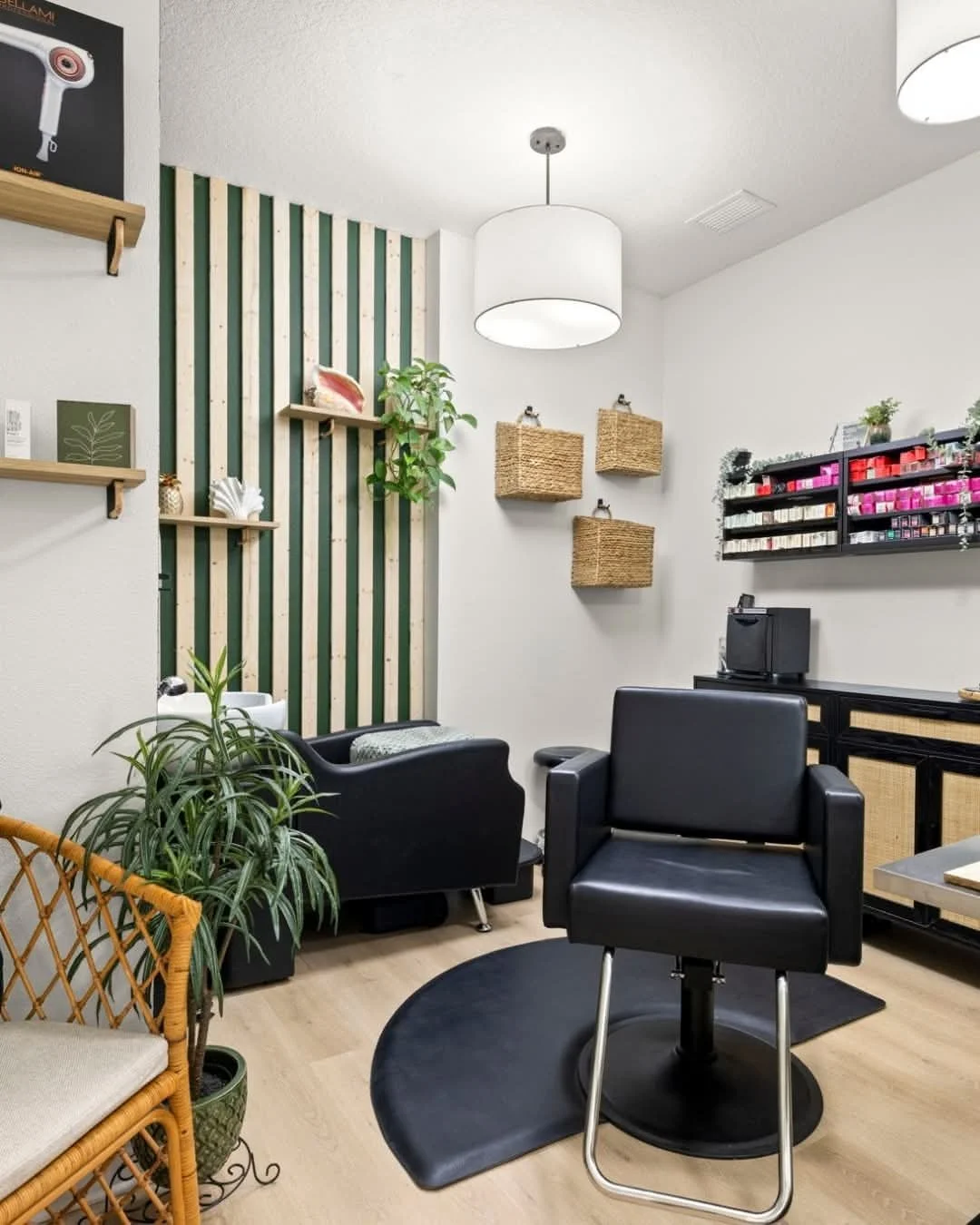 A modern manicure salon with black styling chair, black armchair, potted plant, and decorative wall accents including wooden shelves and woven wall baskets. The space has light wood flooring and a white ceiling with hanging light fixtures.