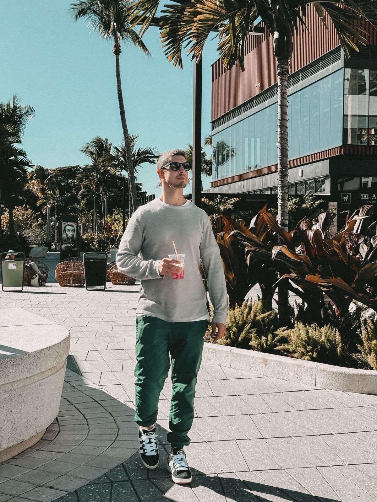 Joven caminando por un área urbana con palmeras, usando gafas de sol, camiseta gris, pantalones verdes y zapatillas, sosteniendo una bebida con popote bajo un cielo azul.