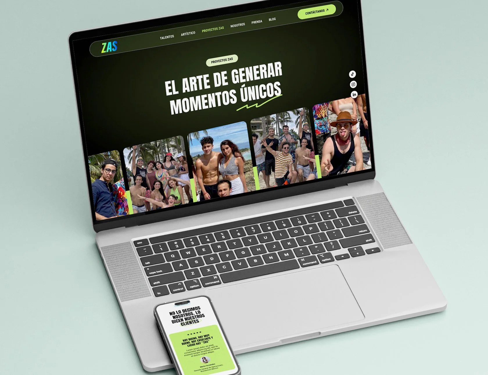 Una computadora portátil y un teléfono móvil en un fondo de color verde menta. La pantalla de la laptop muestra una página web con fotografías de diversas personas en una reunión o evento al aire libre, y un texto que dice 'El arte de generar momentos únicos'.