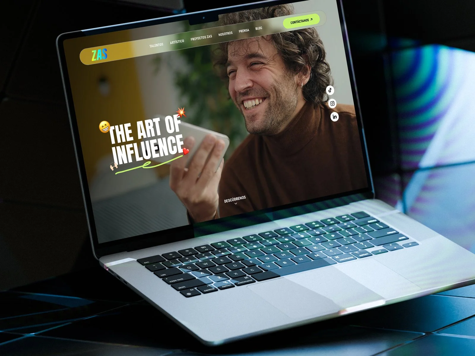Pantalla de laptop mostrando la página web de ZAS con una imagen de un hombre sonriente que sostiene un teléfono móvil, texto que dice 'The Art of Influence' y enlaces a redes sociales.
