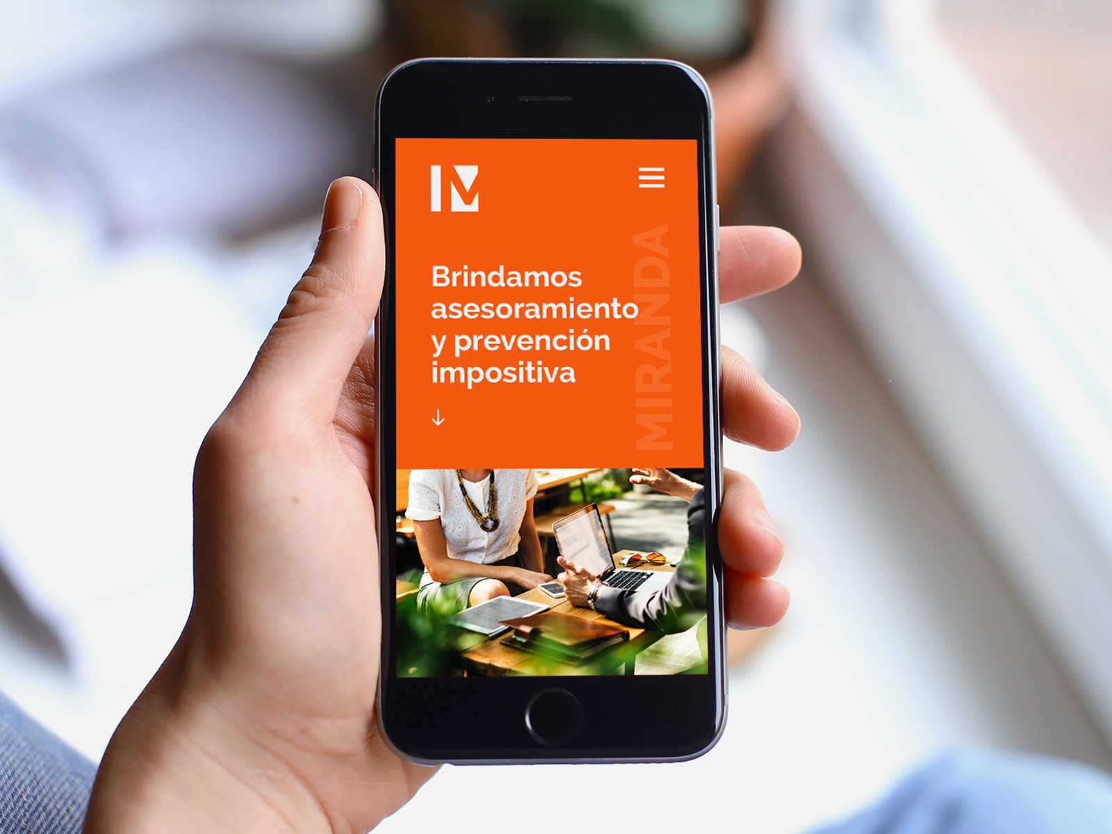 Mano sosteniendo un teléfono móvil que muestra una página web con texto en español y una imagen de dos personas en una reunión de trabajo en un ambiente moderno y natural.