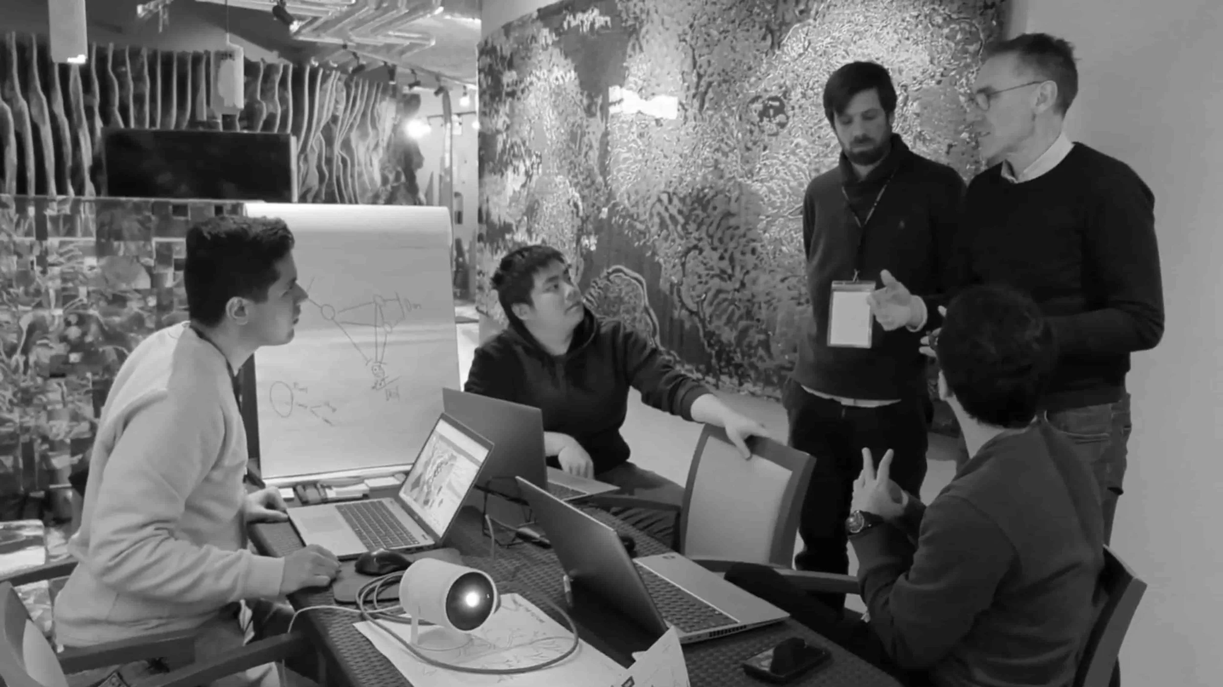 A team of hackers discussing their idea around a table with laptops, a projector, and a flipchart with diagrams.