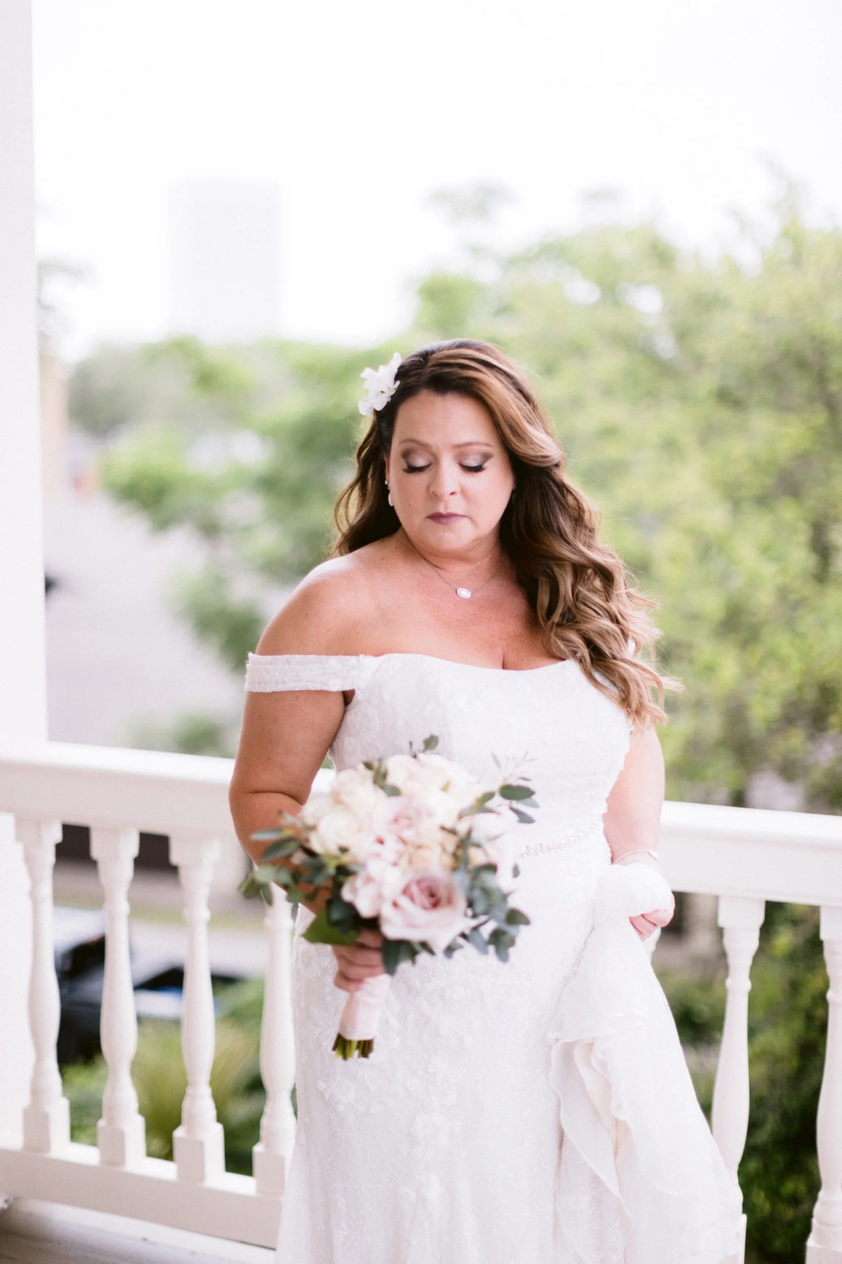 A woman in an off-the-shoulder white wedding dress holding a bouquet of light pink and white flowers, standing outdoors near a white railing with greenery and trees in the background.