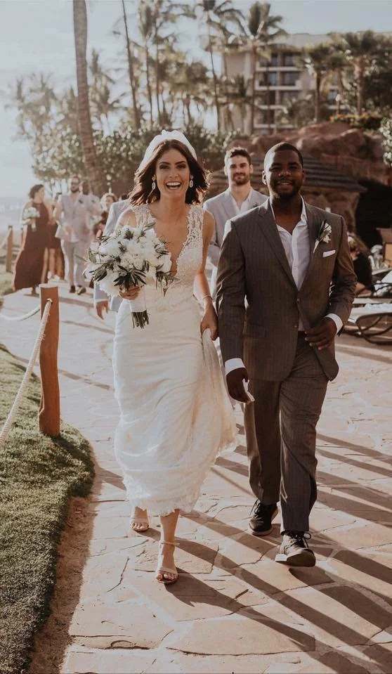A bride in a white wedding dress holding a bouquet walks with a groom in a dark gray suit and white shirt at an outdoor wedding, with friends and palm trees in the background.