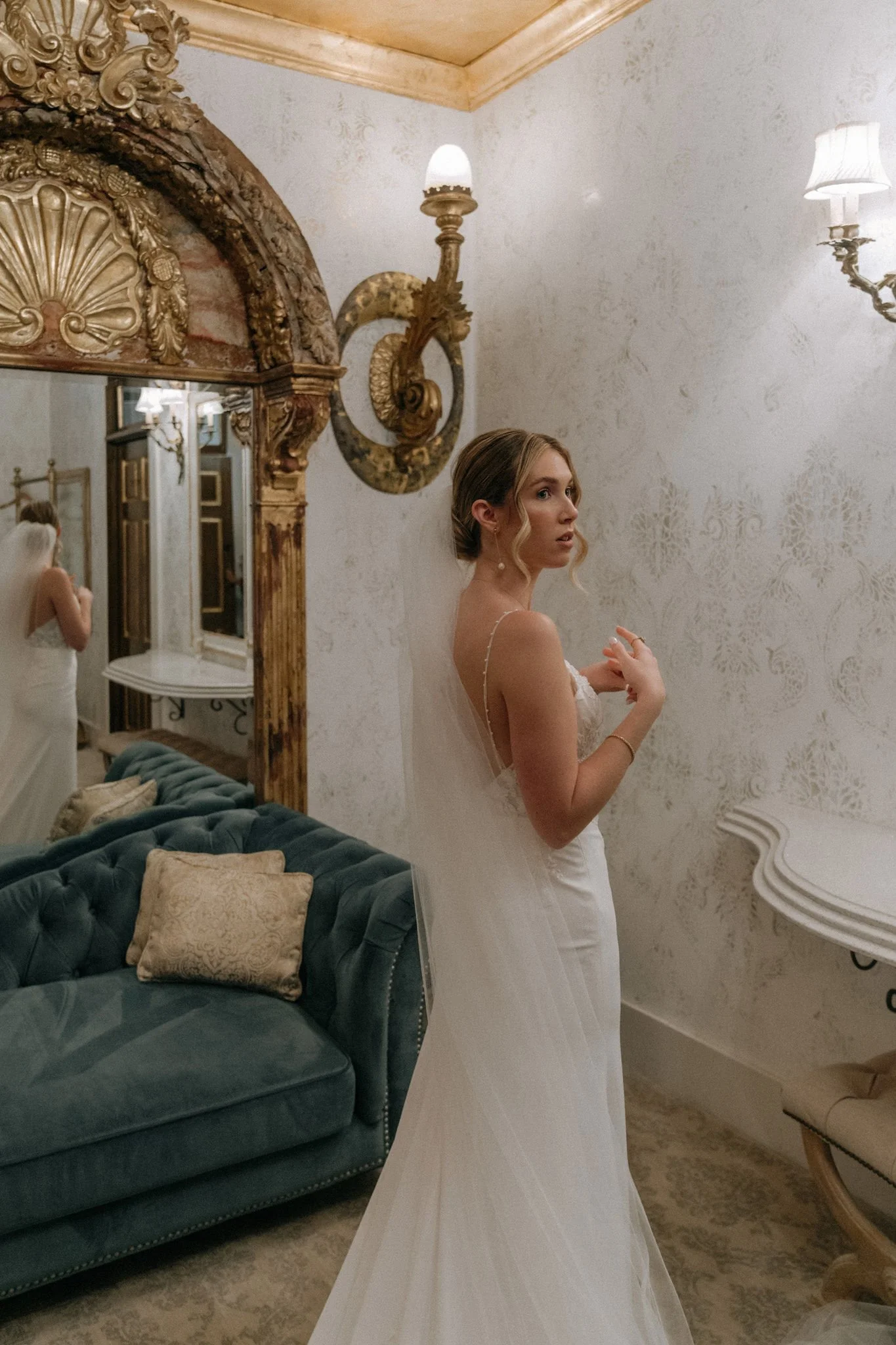 A bride in a white wedding dress standing in a vintage-style room with ornate gold and wood decorations, a teal velvet sofa with decorative pillows, and a mirror reflecting part of the room and another woman in a white dress adjusting her gown.