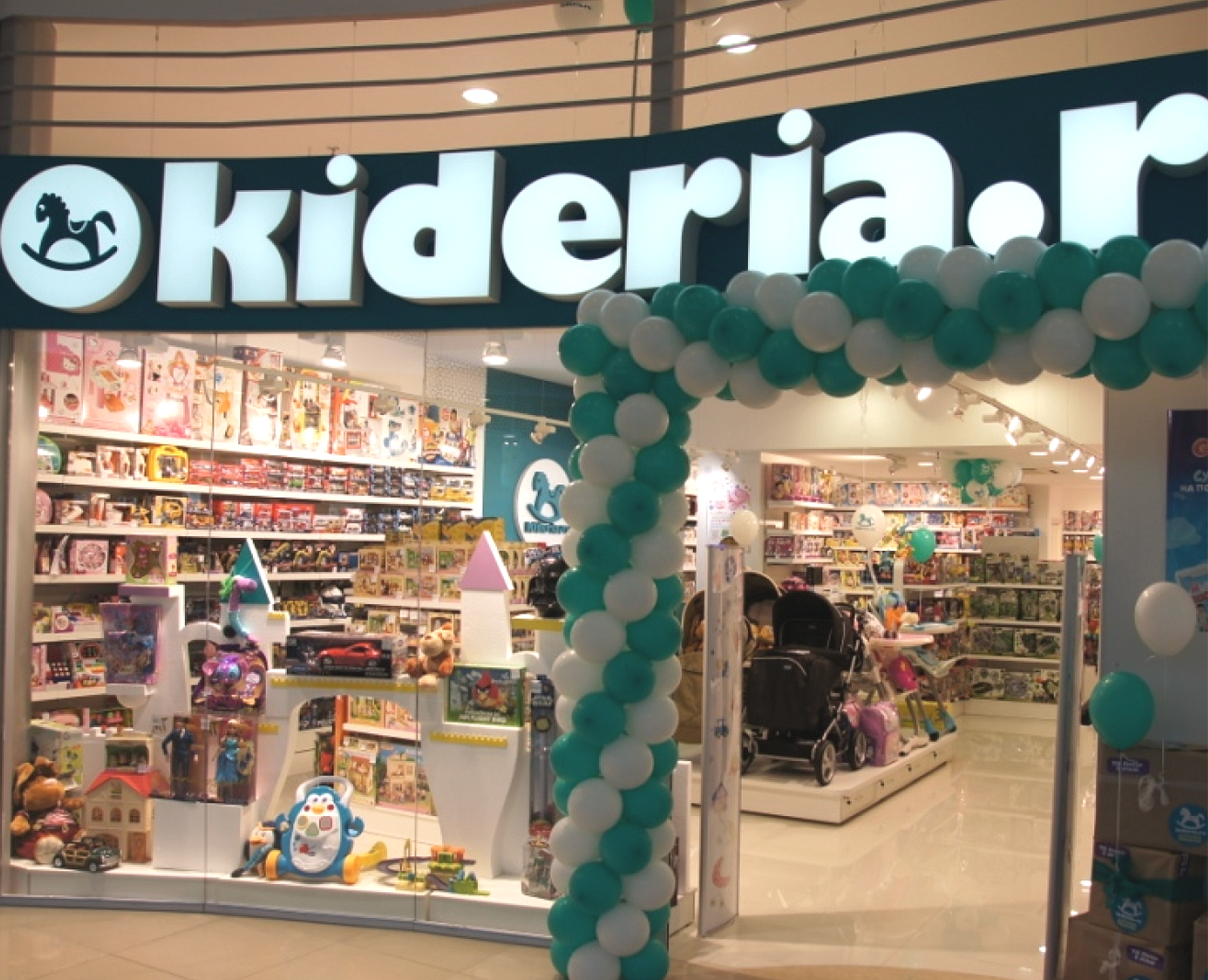 Storefront with illuminated 'Kiderland' sign, decorated with white and teal balloons, displaying children's toys, books, and stuffed animals inside.