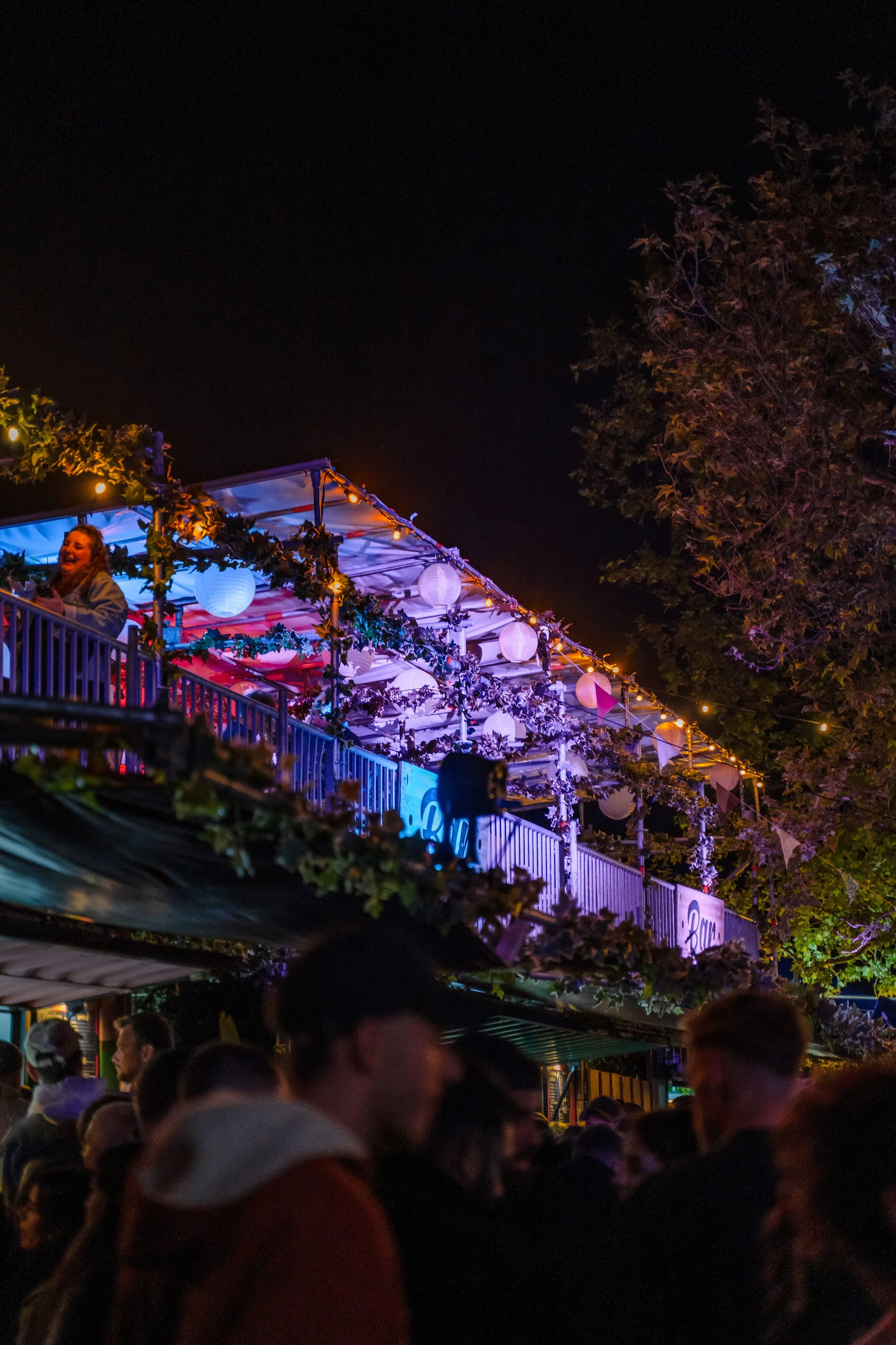 Menschen versammeln sich nachts bei einer festlich beleuchteten Außenveranstaltung mit bunten Lichterketten, Papierlampions und Dekorationen an einer mehrstöckigen Terrasse.