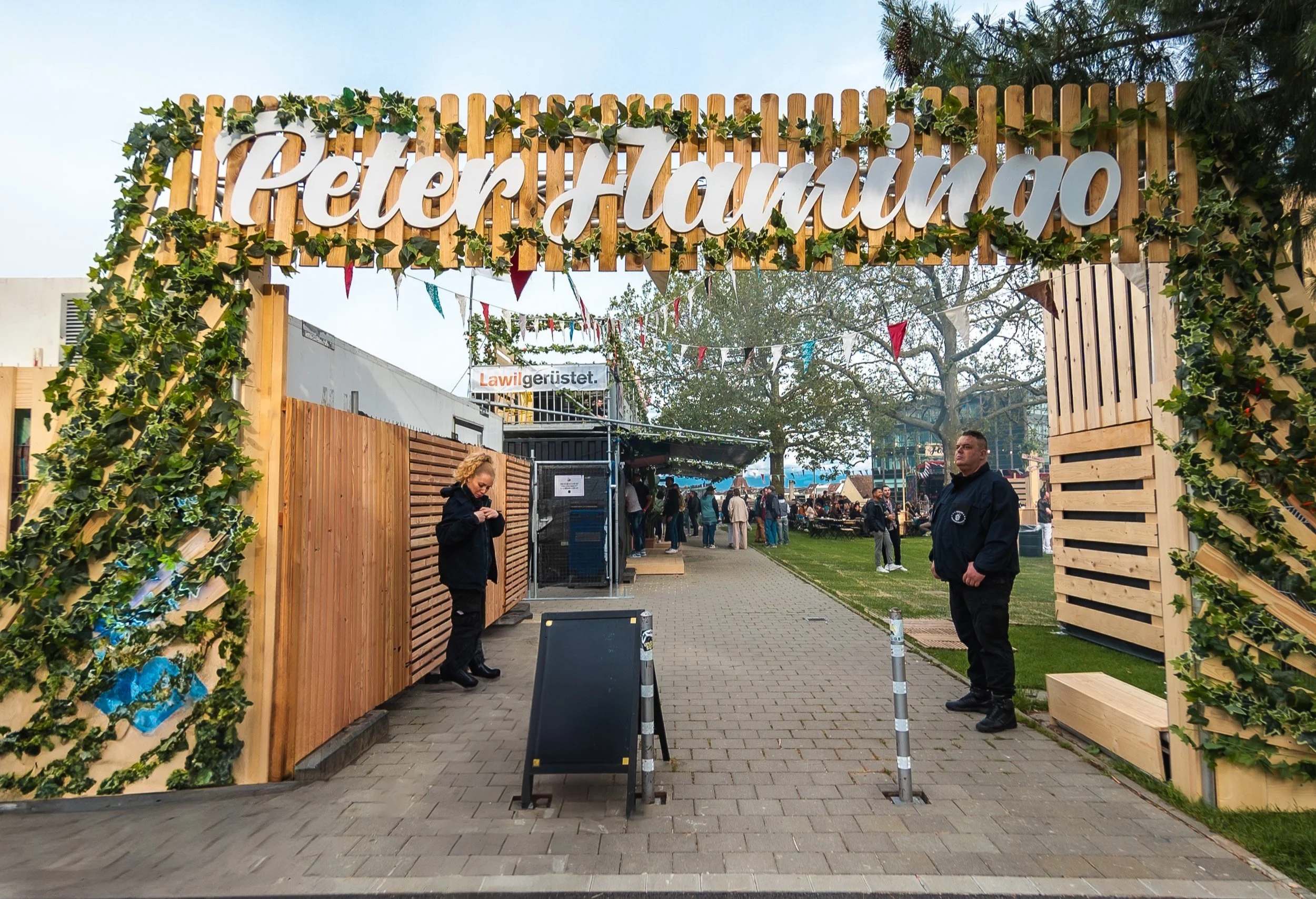 Eingang zu einem Festival namens 'Peter Hammo'. Der Eingang ist mit Holzrahmen und grünen Pflanzen dekoriert. Im Hintergrund sind Menschen, die auf dem Festivalgelände unterwegs sind, und es gibt Bäume und festliche Dekorationen.