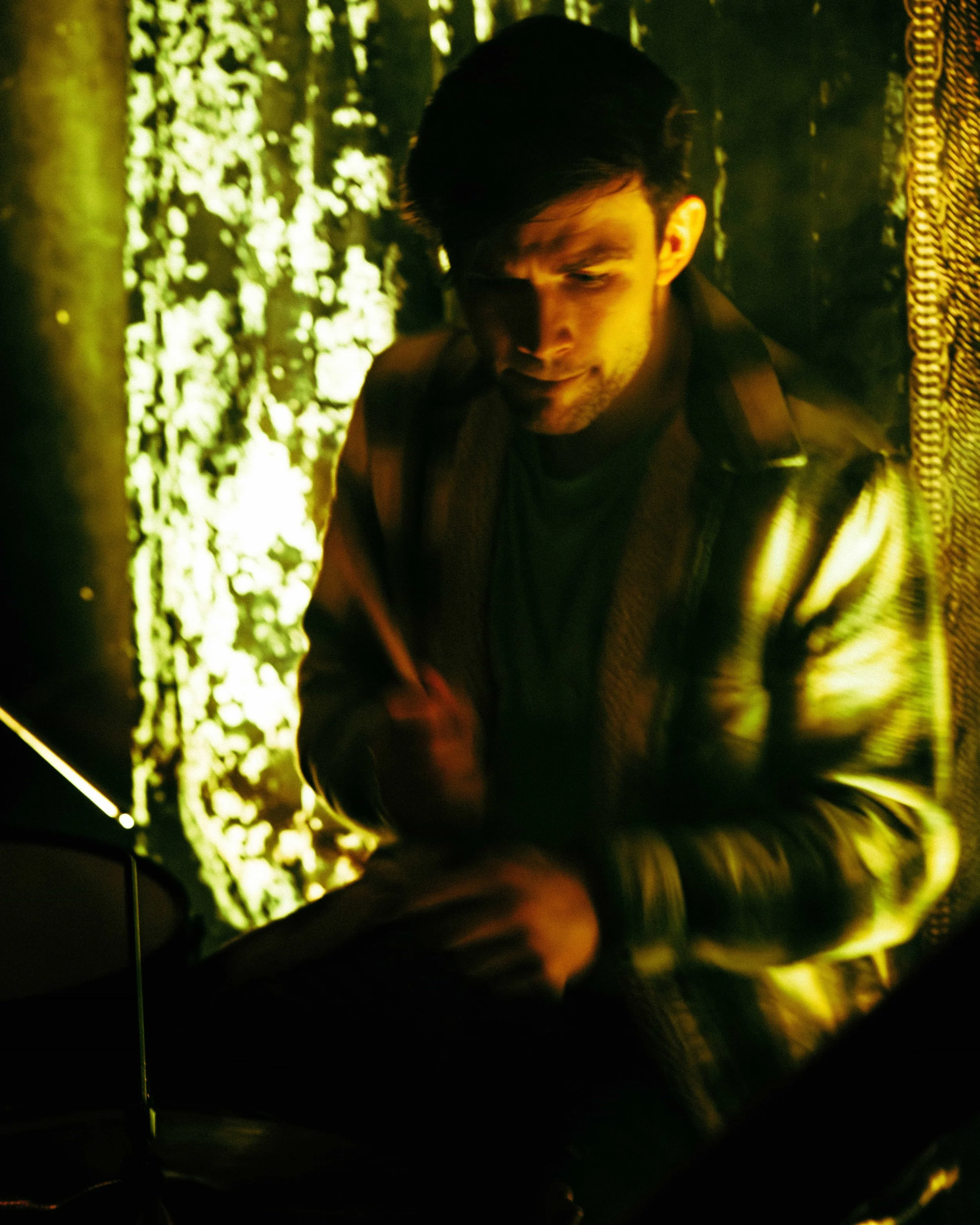 A young man with dark hair and facial hair, sitting in a dimly lit room with yellow-green lighting, holding a phone, with a textured curtain or wall behind him.