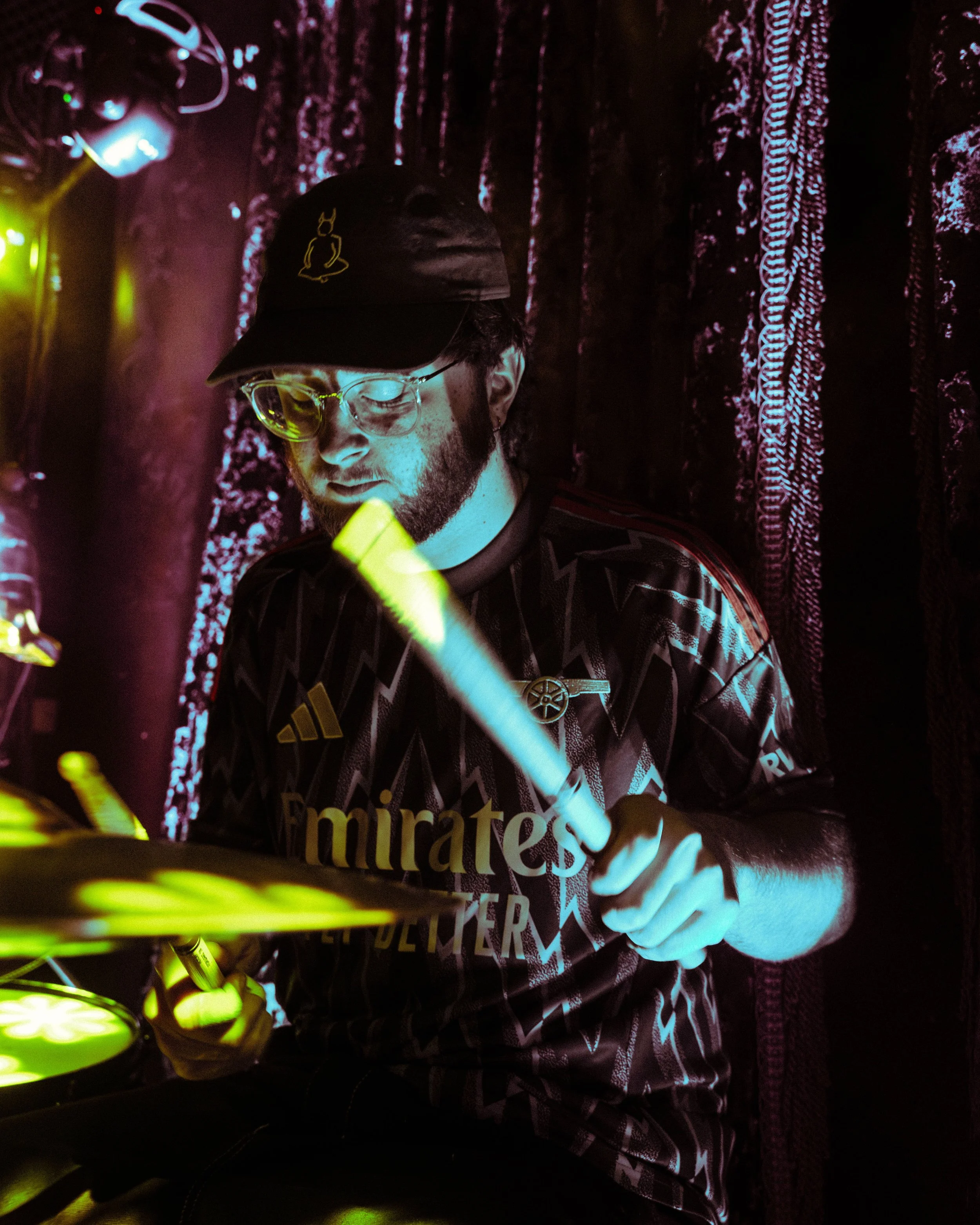 A man with glasses, a baseball cap, and a beard, playing an electronic drum pad under colorful stage lighting.